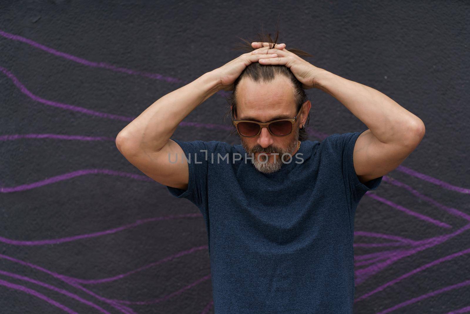 Thoughtful mature man standing with hand folded above his head next to dark urban wall leaned head down wearing sunglasses, dark blue t-shirt and light shorts. Stress, middle age crisis man concept by LipikStockMedia