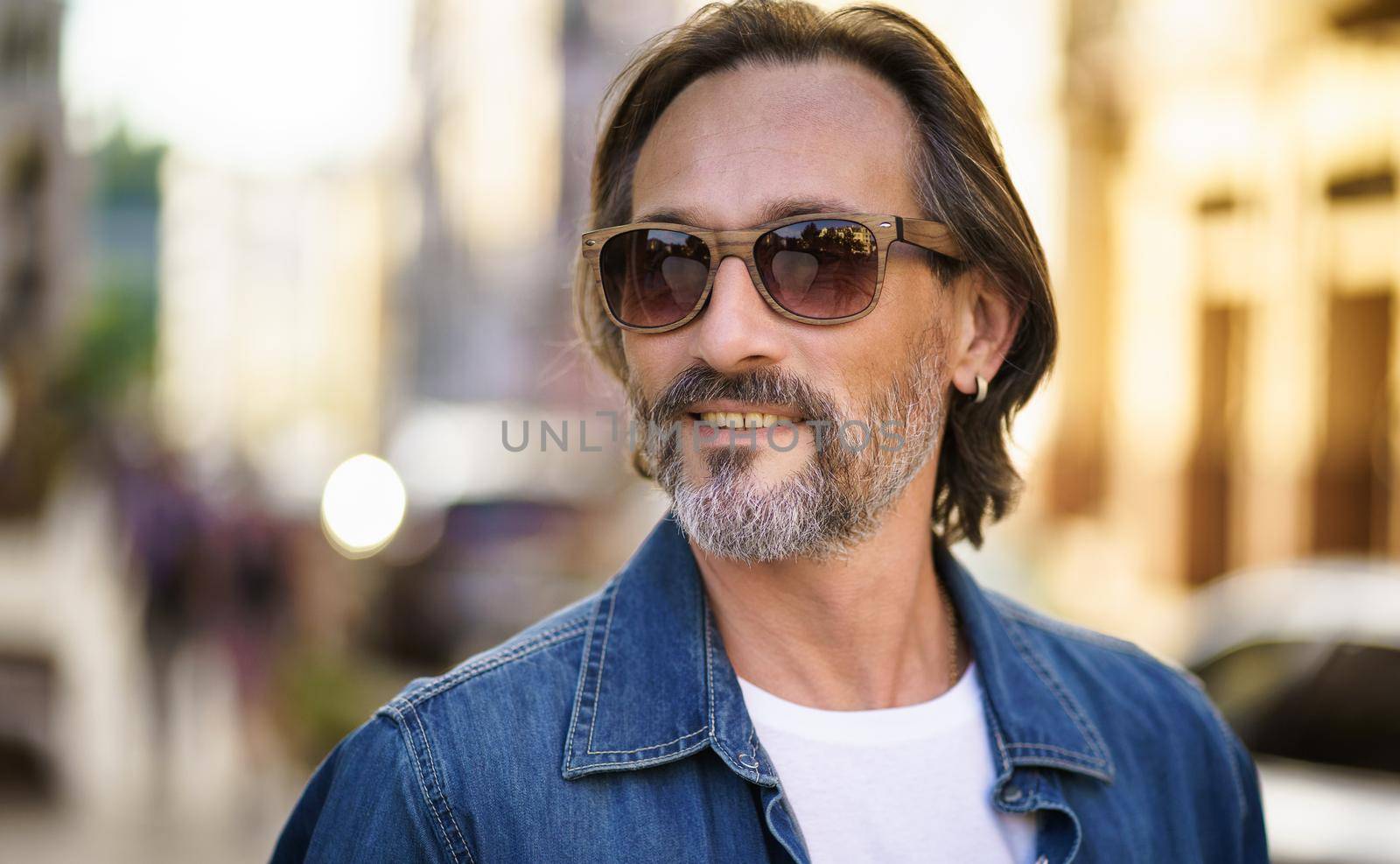 Happy senior man on street of European city wearing sunglasses and denim shirt. Grey bearded Middle aged handsome freelancer man smiling looking sideways wearing casual. Travel concept.