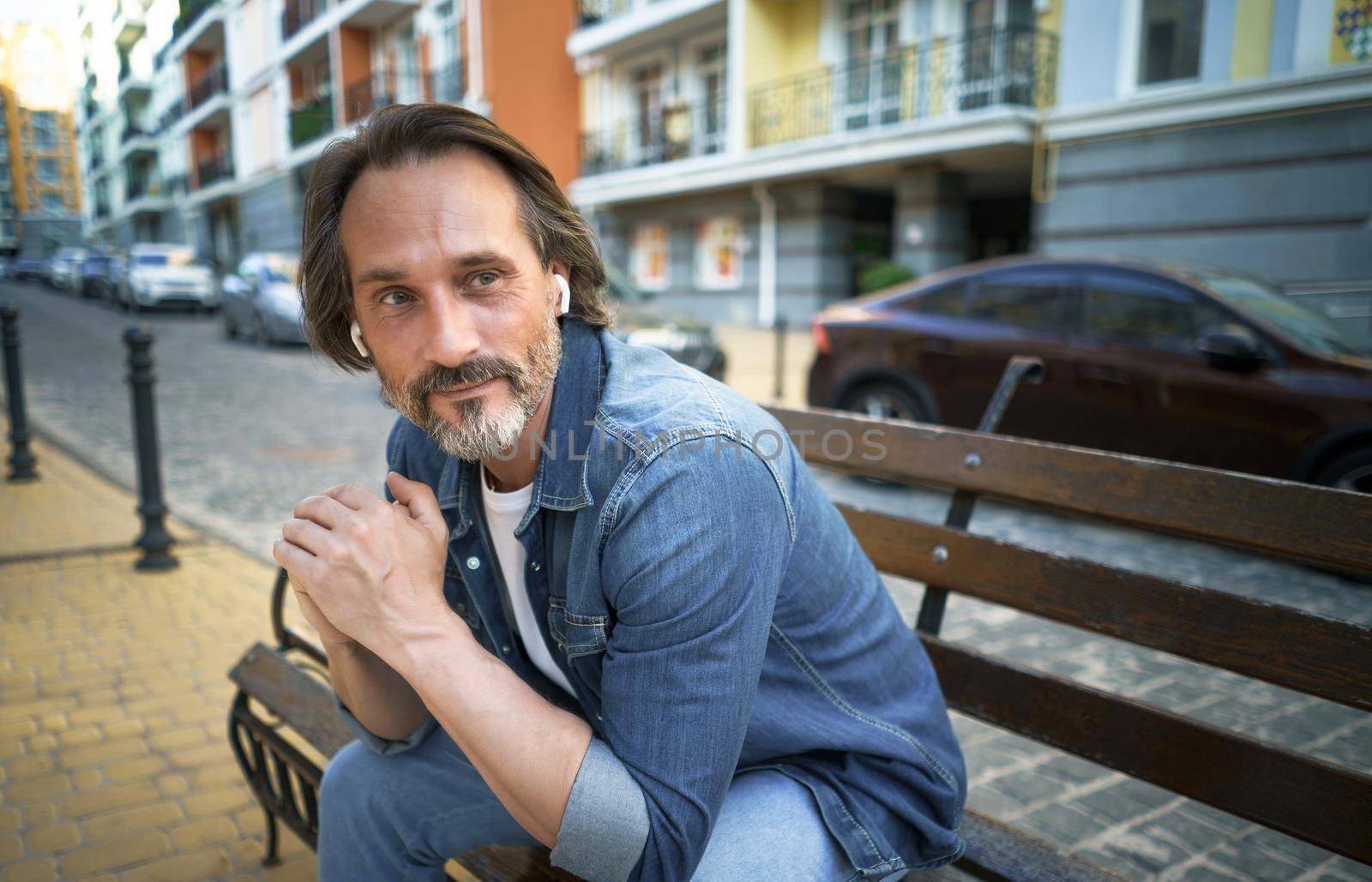 Handsome middle aged man with grey beard spend time talking on the phone or listening music using wireless earphones sitting on the bench in urban city, wearing blue jeans shirt by LipikStockMedia