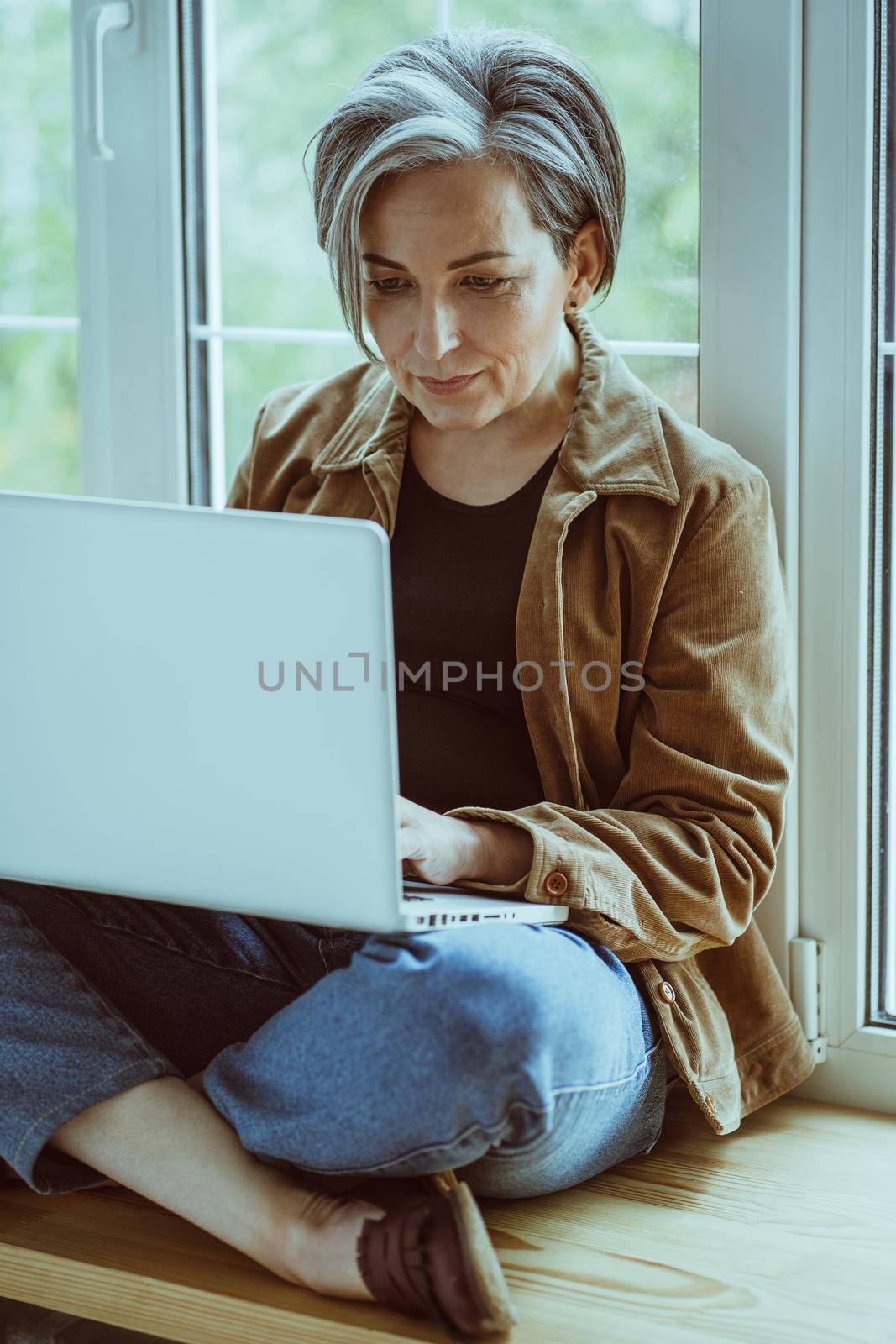 Pretty mature woman works laptop computer sitting on sill while leaning back against the window. Freelance concept. Tinted image.