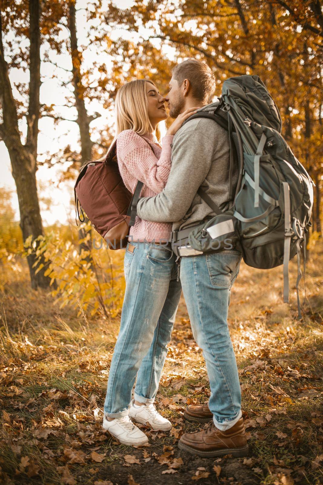 Couple of tourists in love in the autumn forest. Young man and woman kisses and hugs while standing against the backdrop of colorful autumn nature backlit by sunlight. Full-length portrait by LipikStockMedia