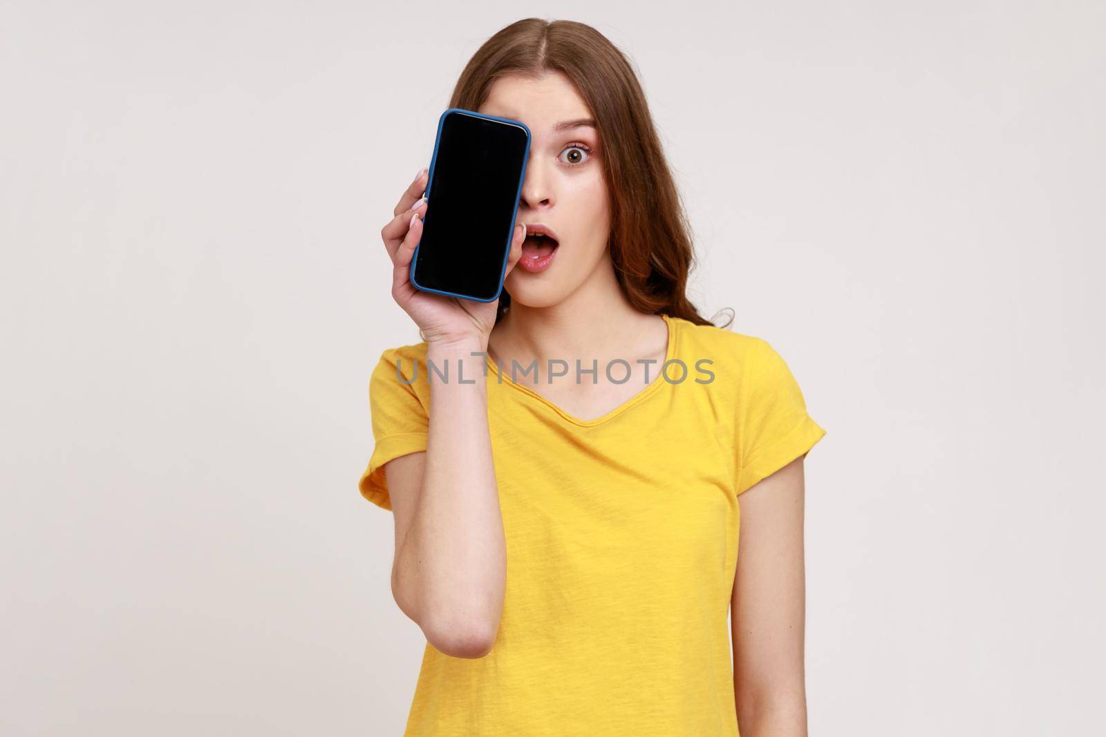 Portrait of excited shocked teenager girl wearing yellow T-shirt covering half face with cell phone and looking at camera with open mouth and big eyes. Indoor studio shot isolated on gray background.