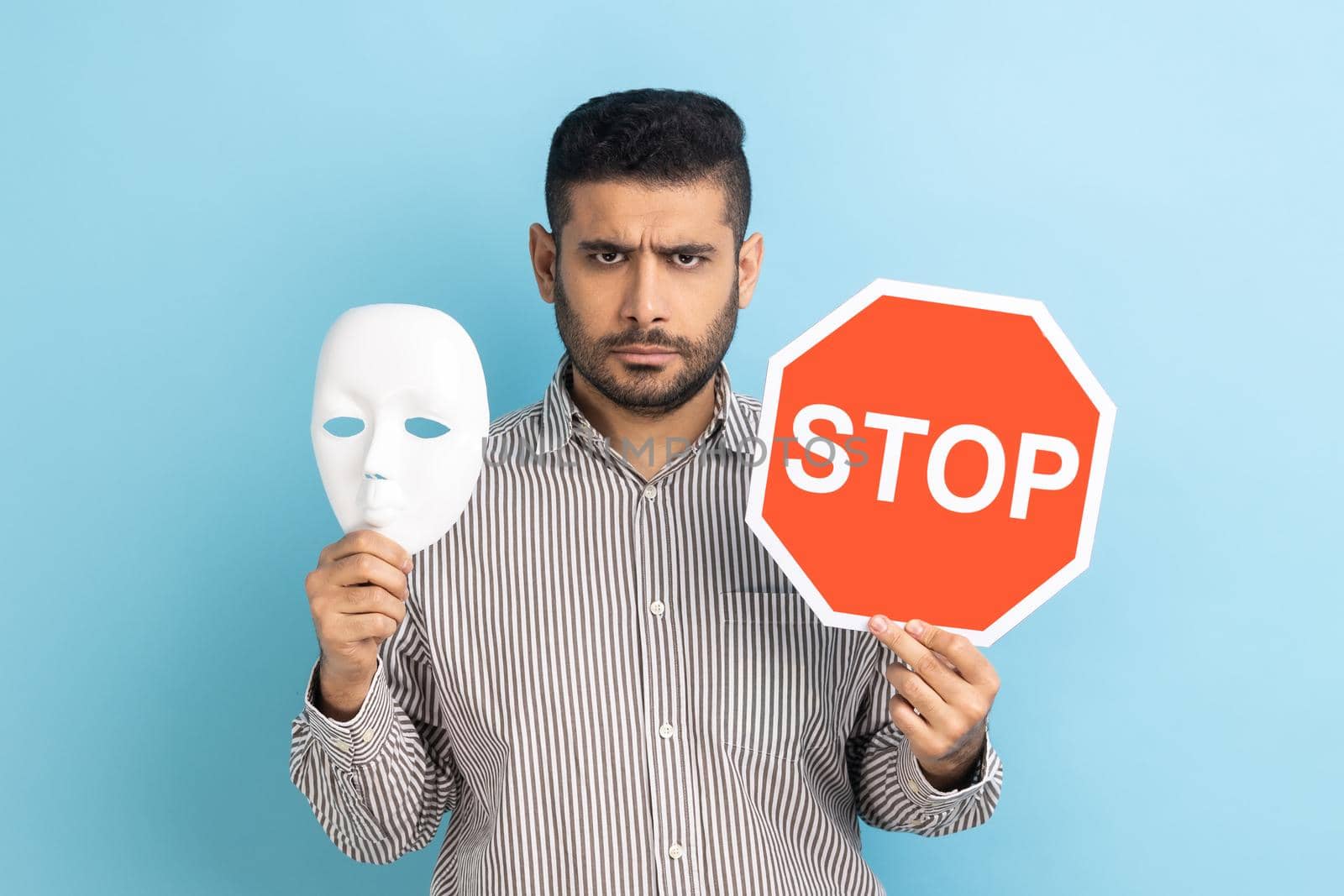 Portrait of serious businessman holding white mask with unknown face and red traffic sign, looking at camera, wearing striped shirt. Indoor studio shot isolated on blue background.