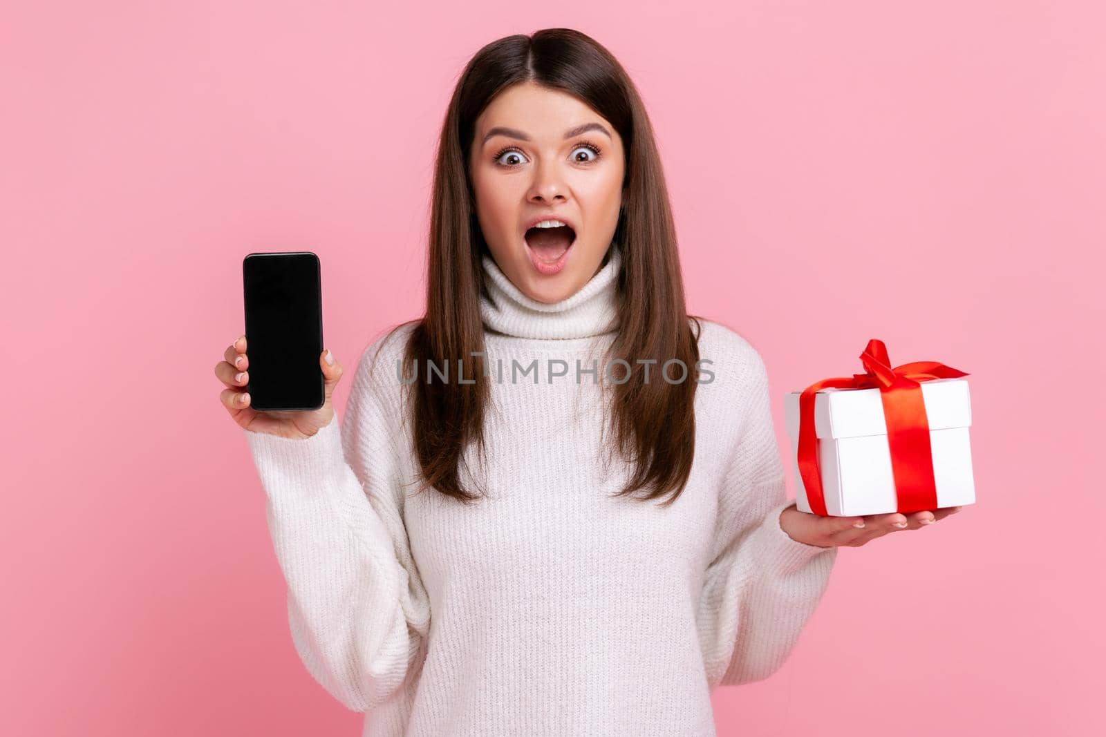 Excited surprised woman holding wrapped present box and cell phone with empty screen for promotion, wearing white casual style sweater. Indoor studio shot isolated on pink background.
