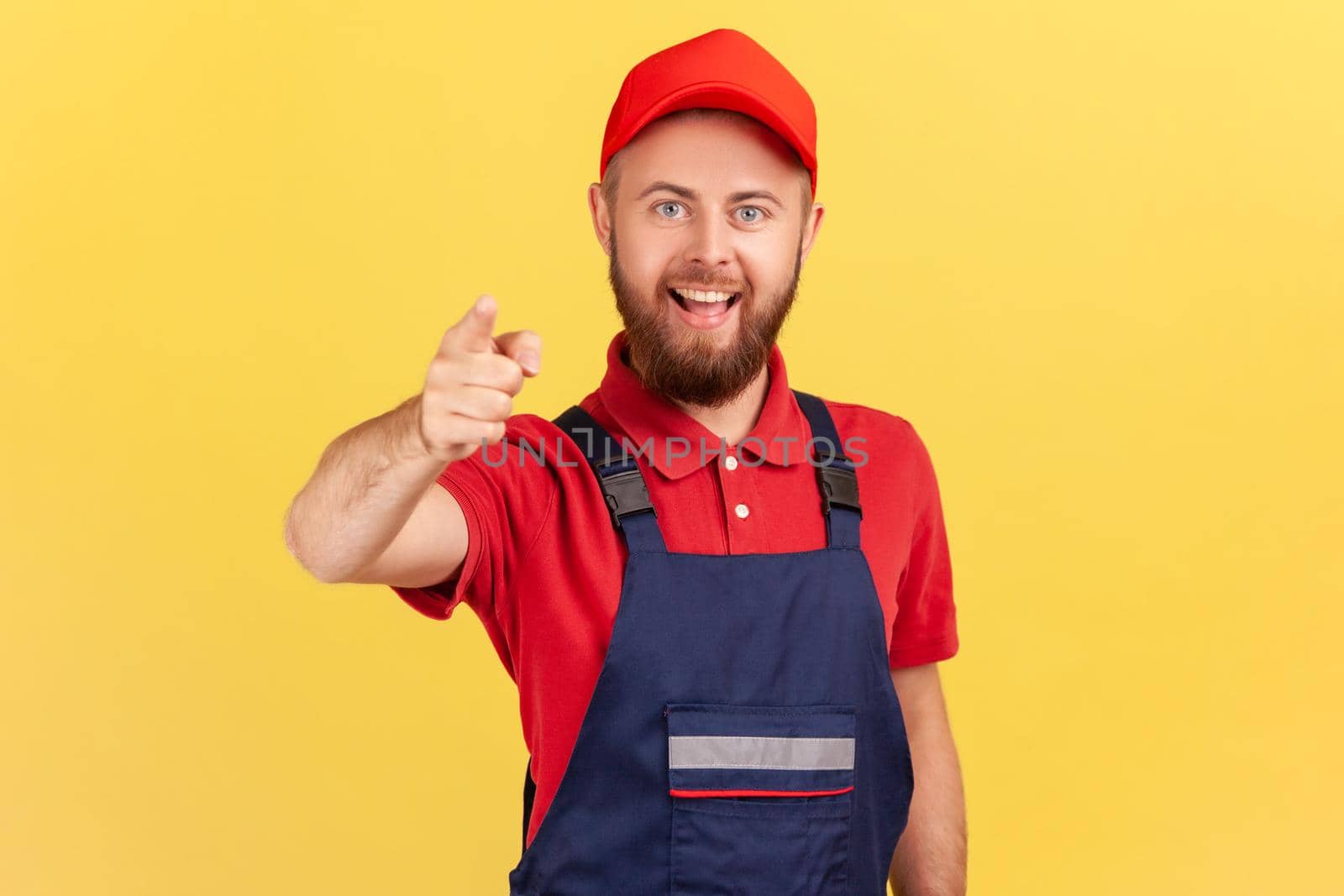 Portrait of smiling bearded satisfied man worker standing and pointing finger to camera, choosing you, wearing overalls and red cap. Indoor studio shot isolated on yellow background.