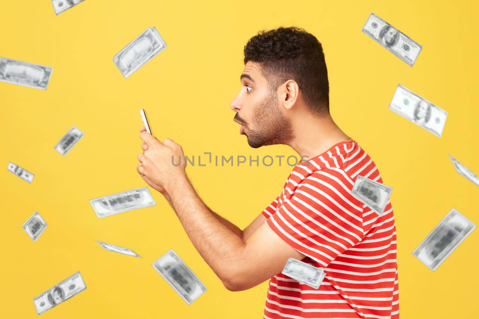 Profile side view of money rain and rich surprised shocked man man looking at smartphone screen with shocked face and can not believe. Indoor studio shot isolated on yellow background