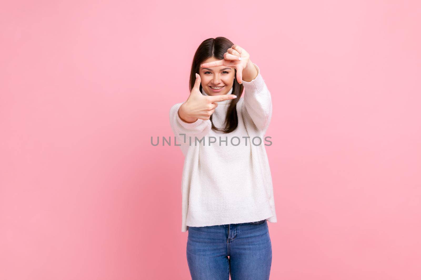 Smiling woman looking through finger frame, imitating to take photo, focusing interesting moment, wearing white casual style sweater. Indoor studio shot isolated on pink background.