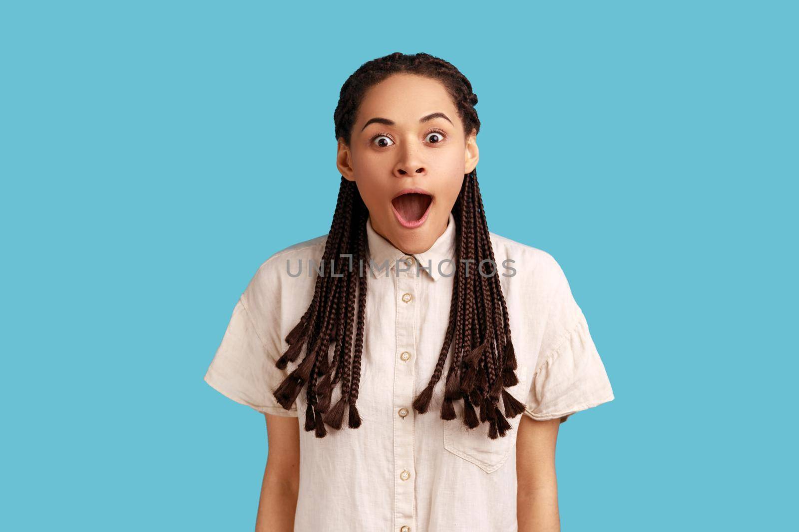 Happy surprised beautiful woman with black dreadlocks notices something incredible, stares amazed, keeps mouth opened, wearing white shirt. Indoor studio shot isolated on blue background.