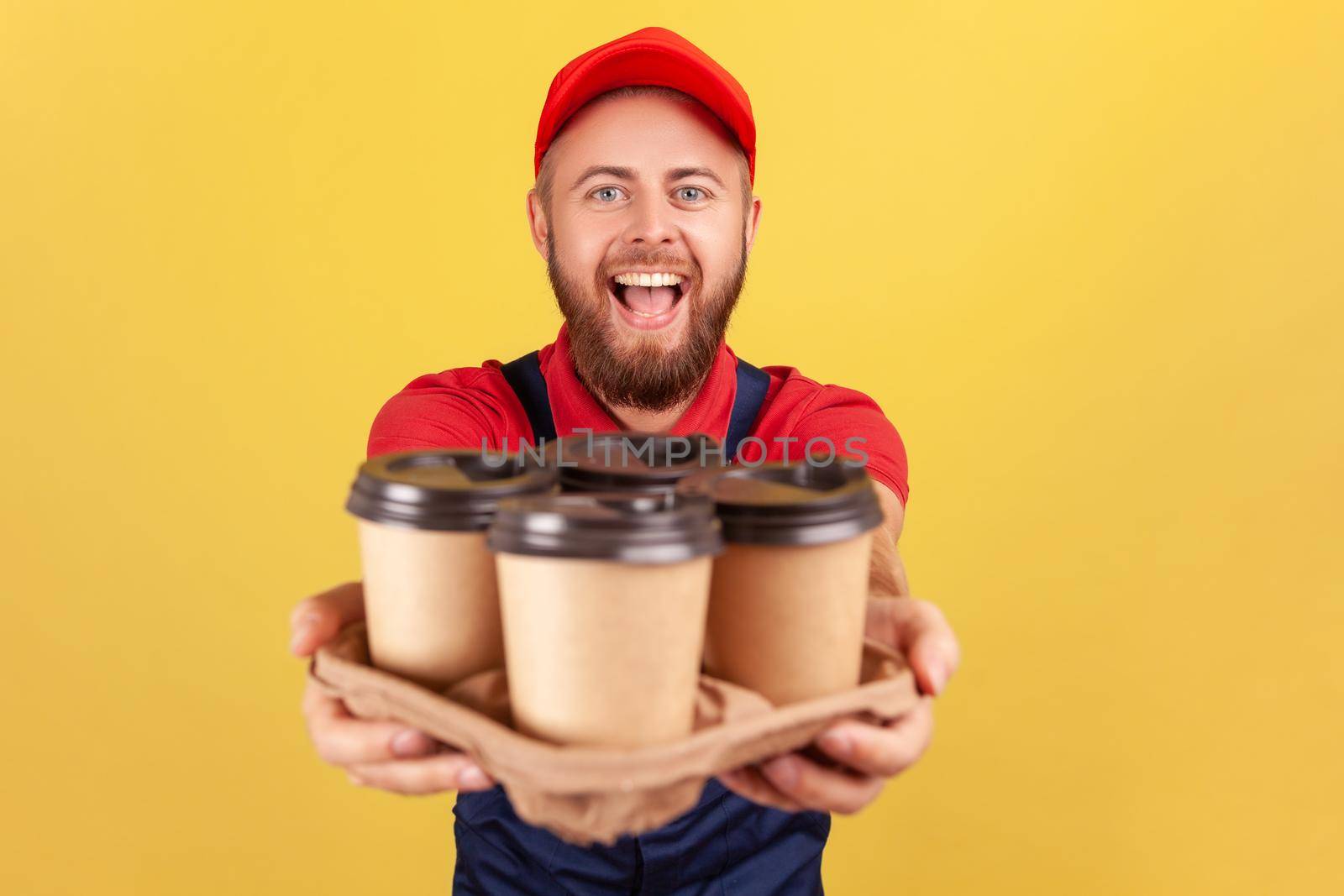 Delivery service. Friendly happy courier man in blue overalls offering coffee, giving drinks in disposable cups, looking at camera with excited look. Indoor studio shot isolated on yellow background.