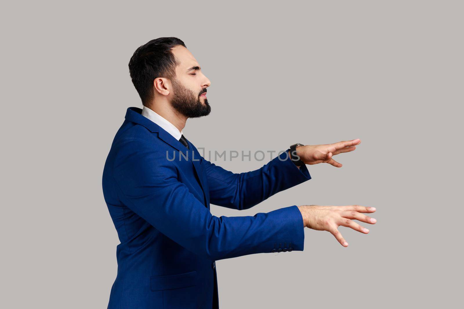 Side view of sick blind bearded businessman walking with eyes closed and holding out his hands to find road, wearing official style suit. Indoor studio shot isolated on gray background.