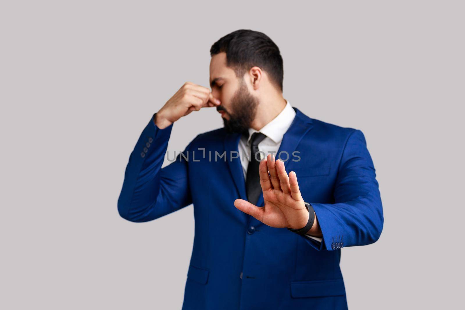 Bearded man grabbing nose, grimacing in disgust and showing stop gesture, expressing repulsion to stink, fart gases, wearing official style suit. Indoor studio shot isolated on gray background.