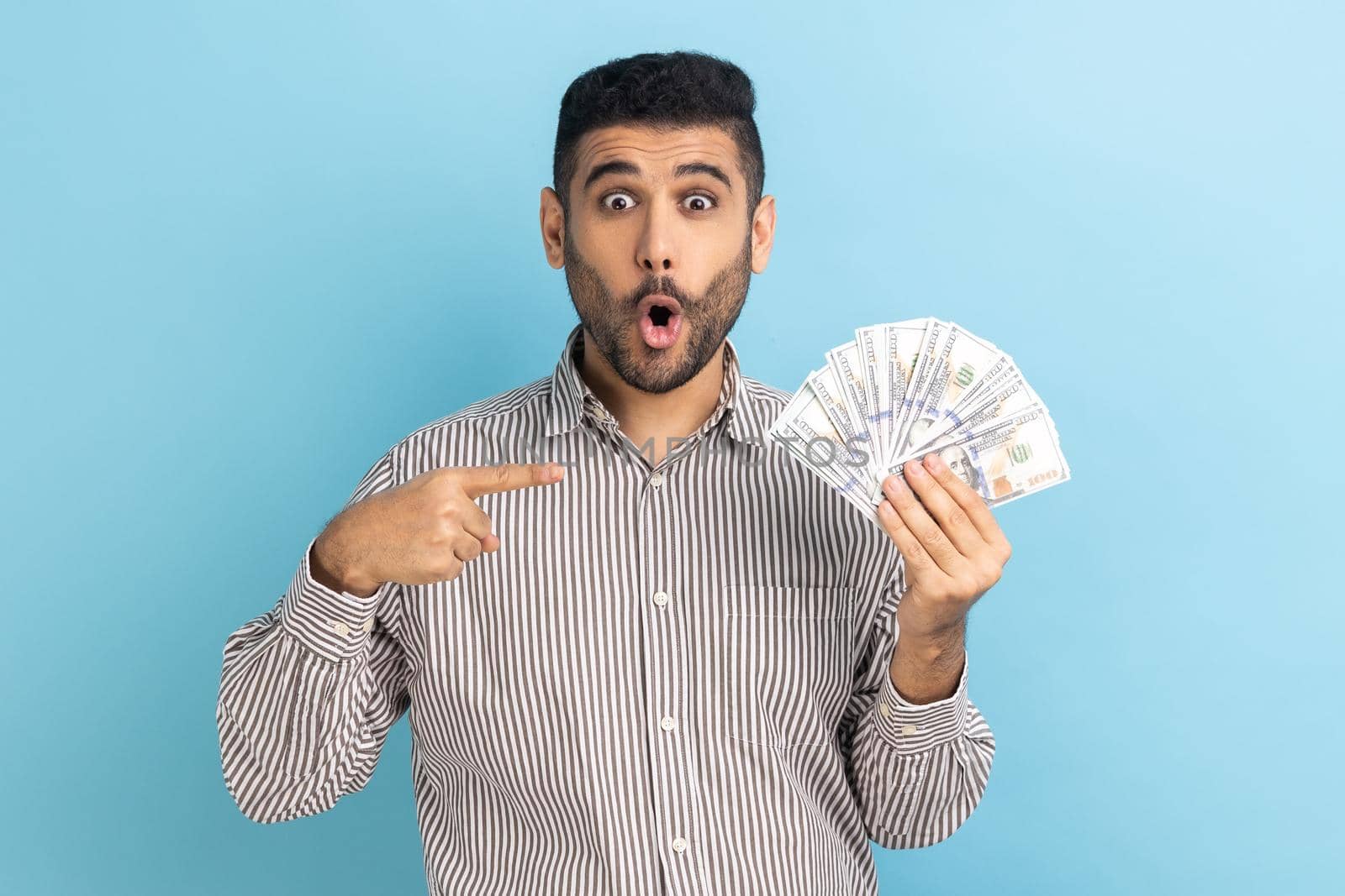 Shocked businessman holding lot of money, pointing at dollar bills in hand and keeps mouth open, excited of lottery win, wearing striped shirt. Indoor studio shot isolated on blue background.