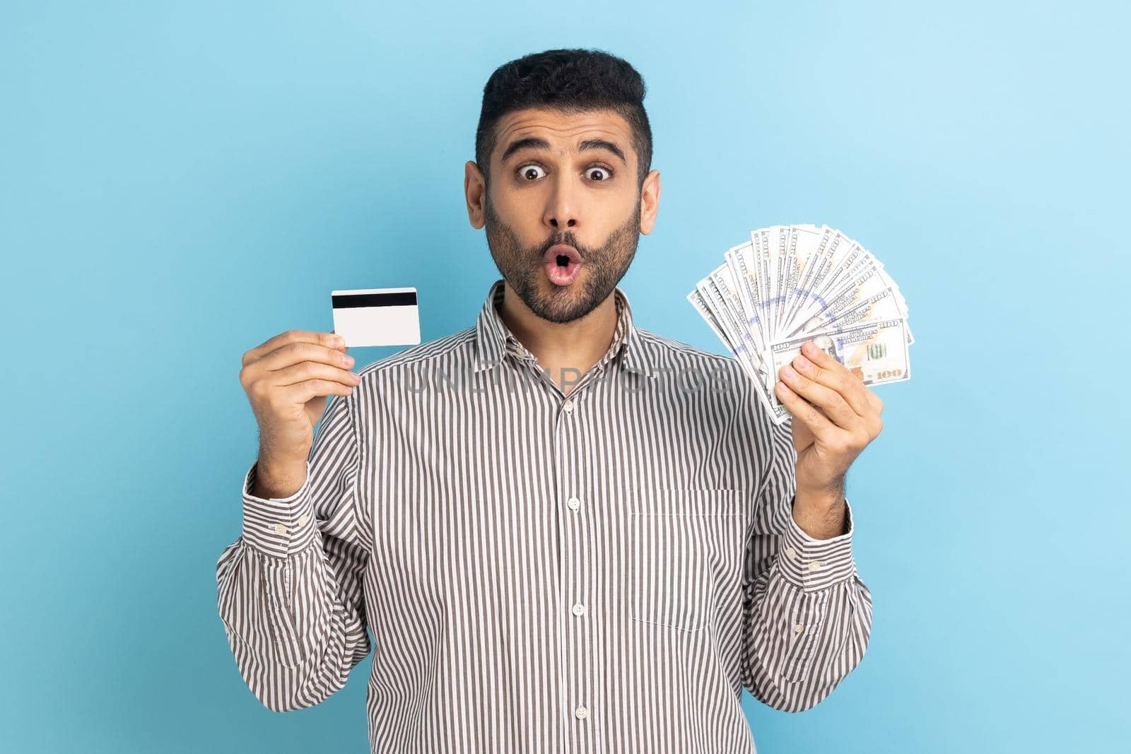 Astonished businessman with beard holding dollar banknotes and credit card, looking at camera with open mouth, wearing striped shirt. Indoor studio shot isolated on blue background.