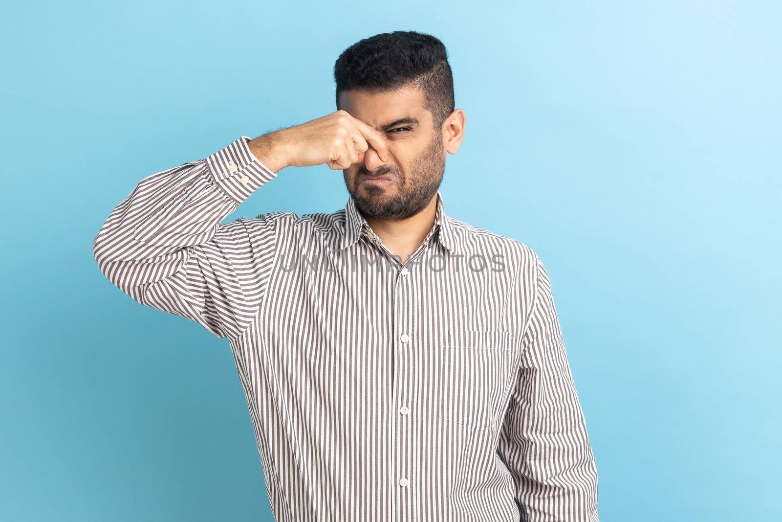 Portrait of young confused businessman standing pinching his nose and looking at camera because back smell, wearing striped shirt. Indoor studio shot isolated on blue background.