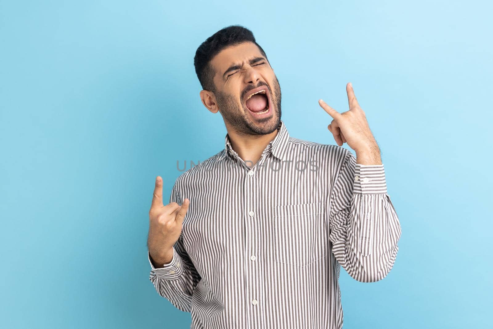 Disobedient businessman with beard showing rock and roll gesture and excitedly screaming, having fun, enjoying heavy music, wearing striped shirt. Indoor studio shot isolated on blue background.