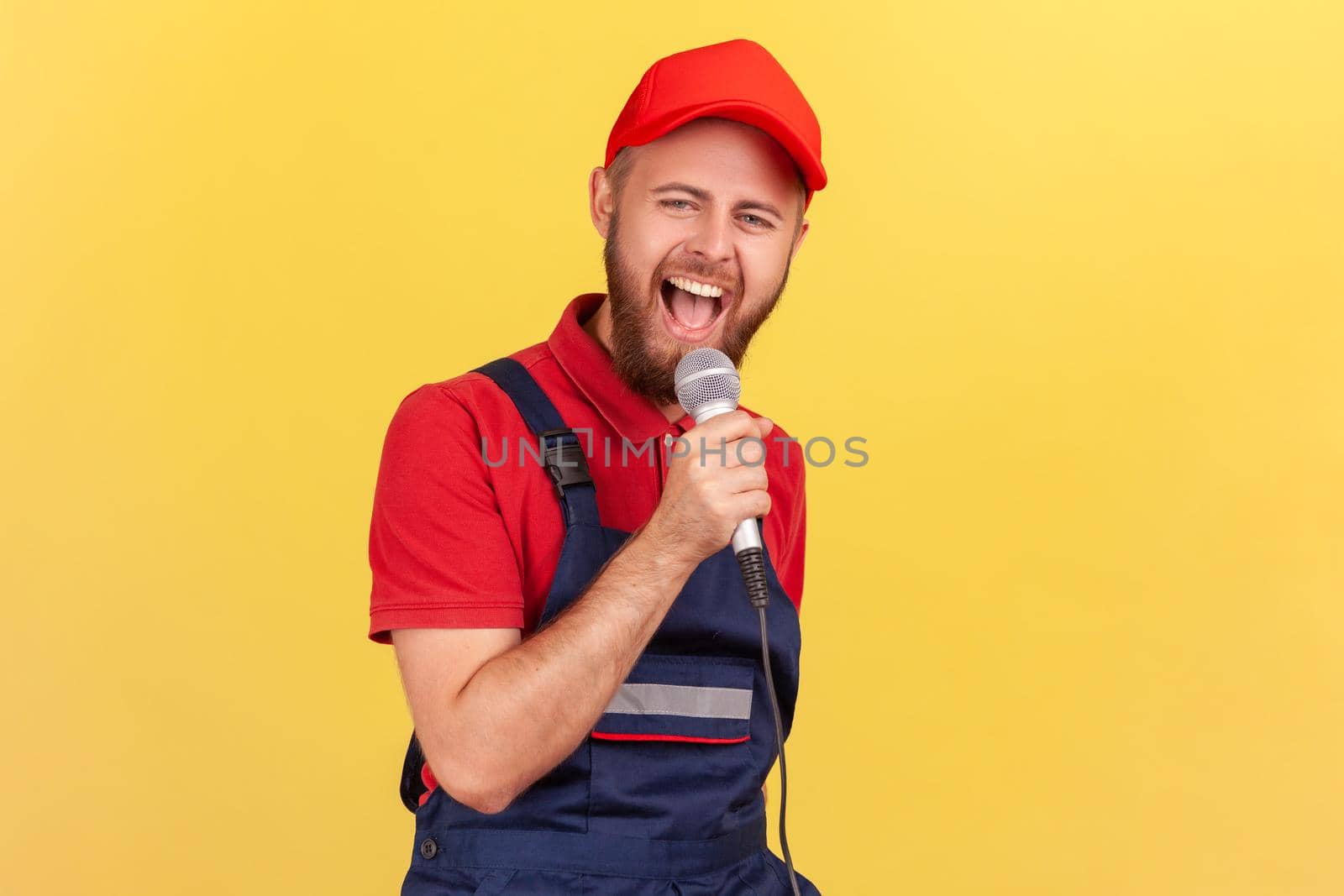Portrait of happy excited worker man standing with microphone in hands and singing at corporate party, having fun at work with colleagues. Indoor studio shot isolated on yellow background.
