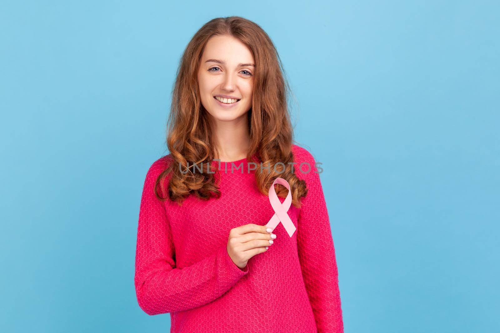Lovely beautiful woman wearing pink pullover, holding pink ribbon, breast cancer awareness, female health diagnostics and cancer prevention. Indoor studio shot isolated on blue background.