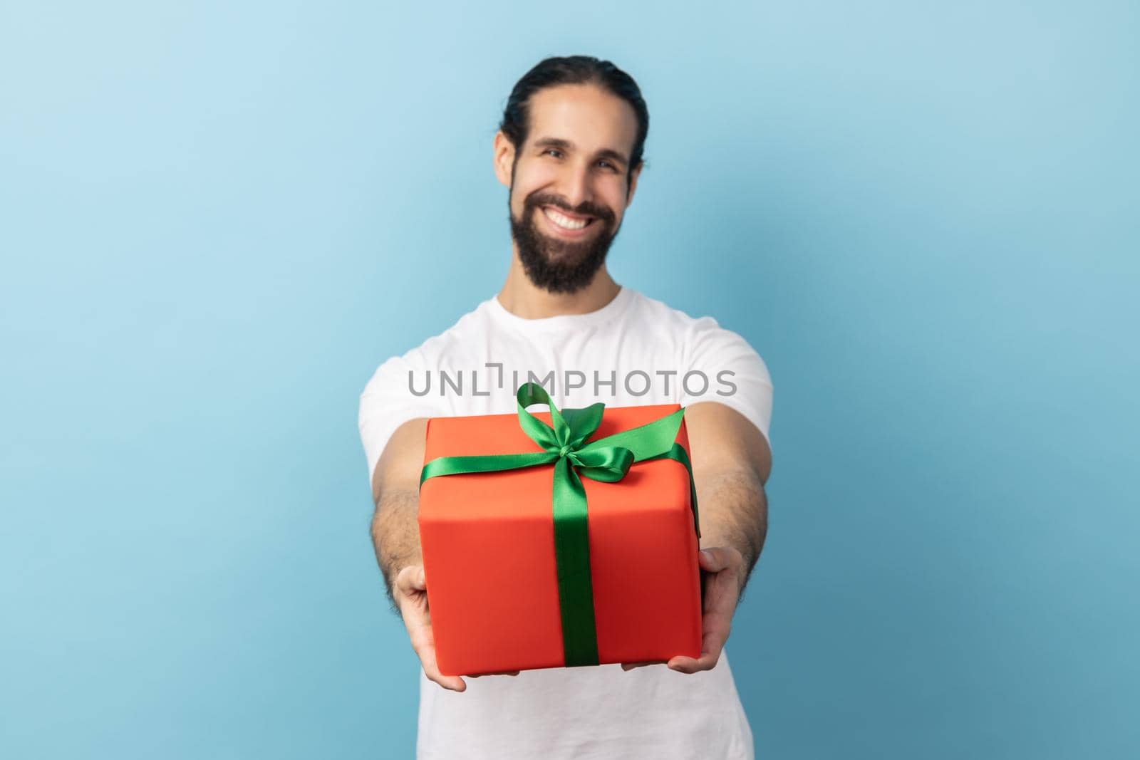 Portrait of handsome delighted man with beard wearing white T-shirt giving wrapped gift box and smiling at camera, happy holiday. Indoor studio shot isolated on blue background.