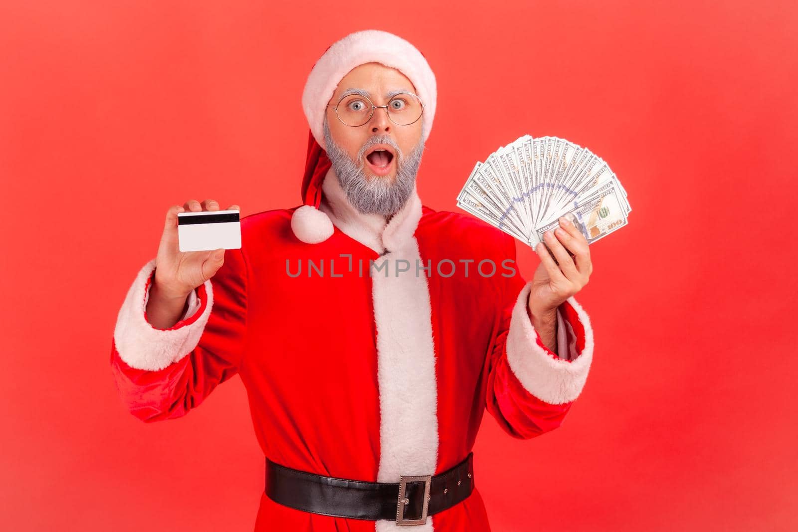 Portrait of shocked elderly man with gray beard wearing santa claus costume holding fan of dollars and credit card, astonished of big cashback. Indoor studio shot isolated on red background.