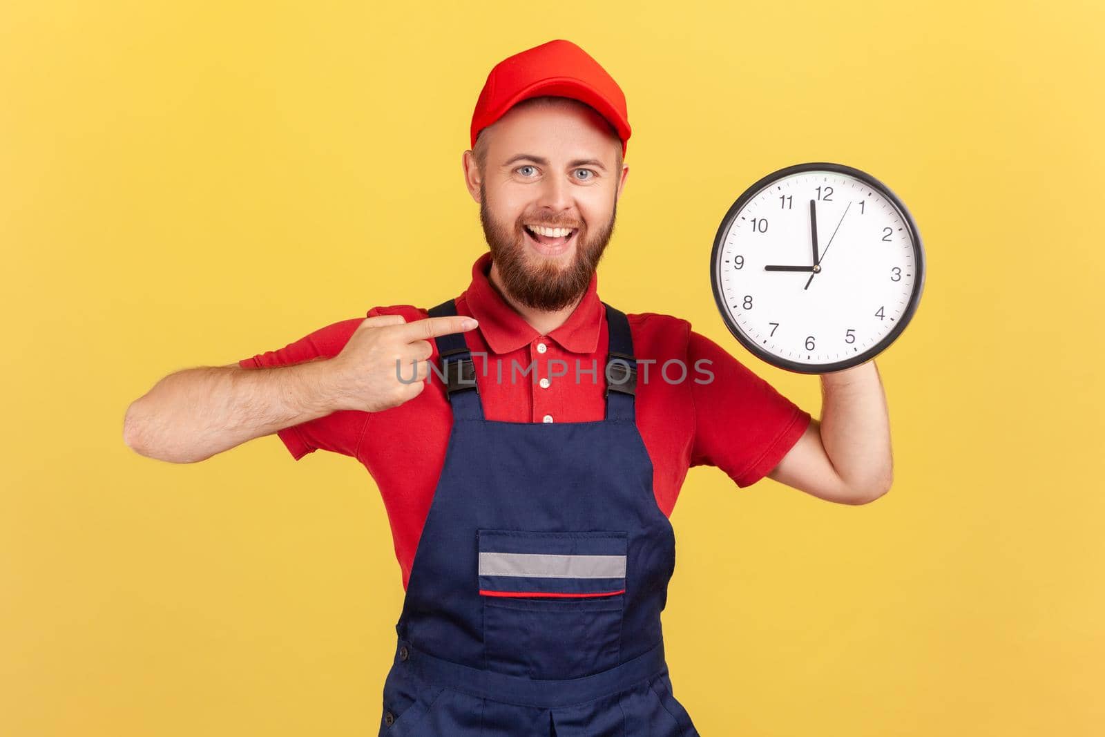 Portrait of delighted man worker standing and pointing at big wall clock in his hands, looking at camera, wearing overalls and red cap. Indoor studio shot isolated on yellow background.