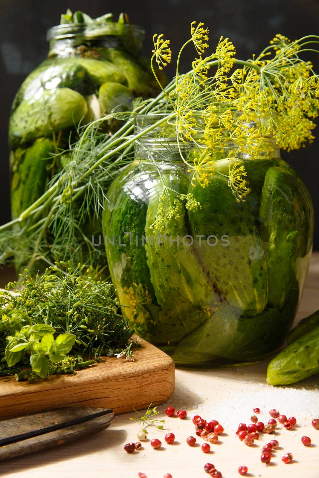 Two jars with home made pickled cucumbers with dill and other herbs and spices on white wooden table, selective focus.