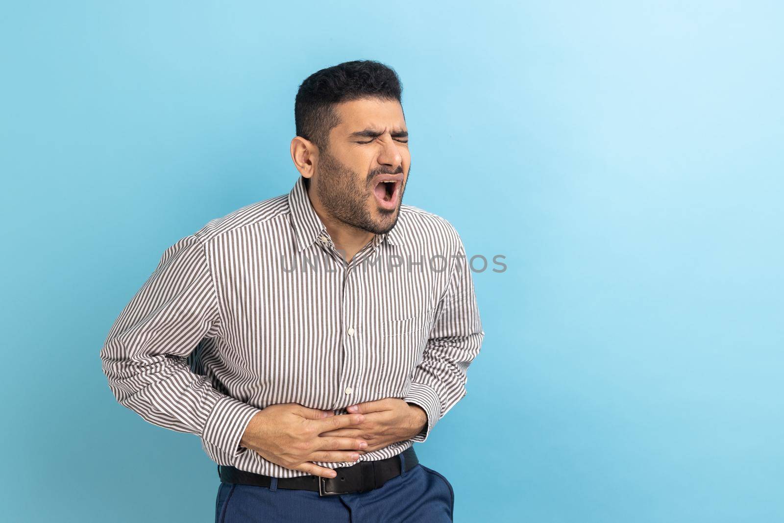 Portrait of sick bearded businessman hunching and grimacing from strong stomach ache, having indigestion or diarrhea symptoms, wearing striped shirt. Indoor studio shot isolated on blue background.