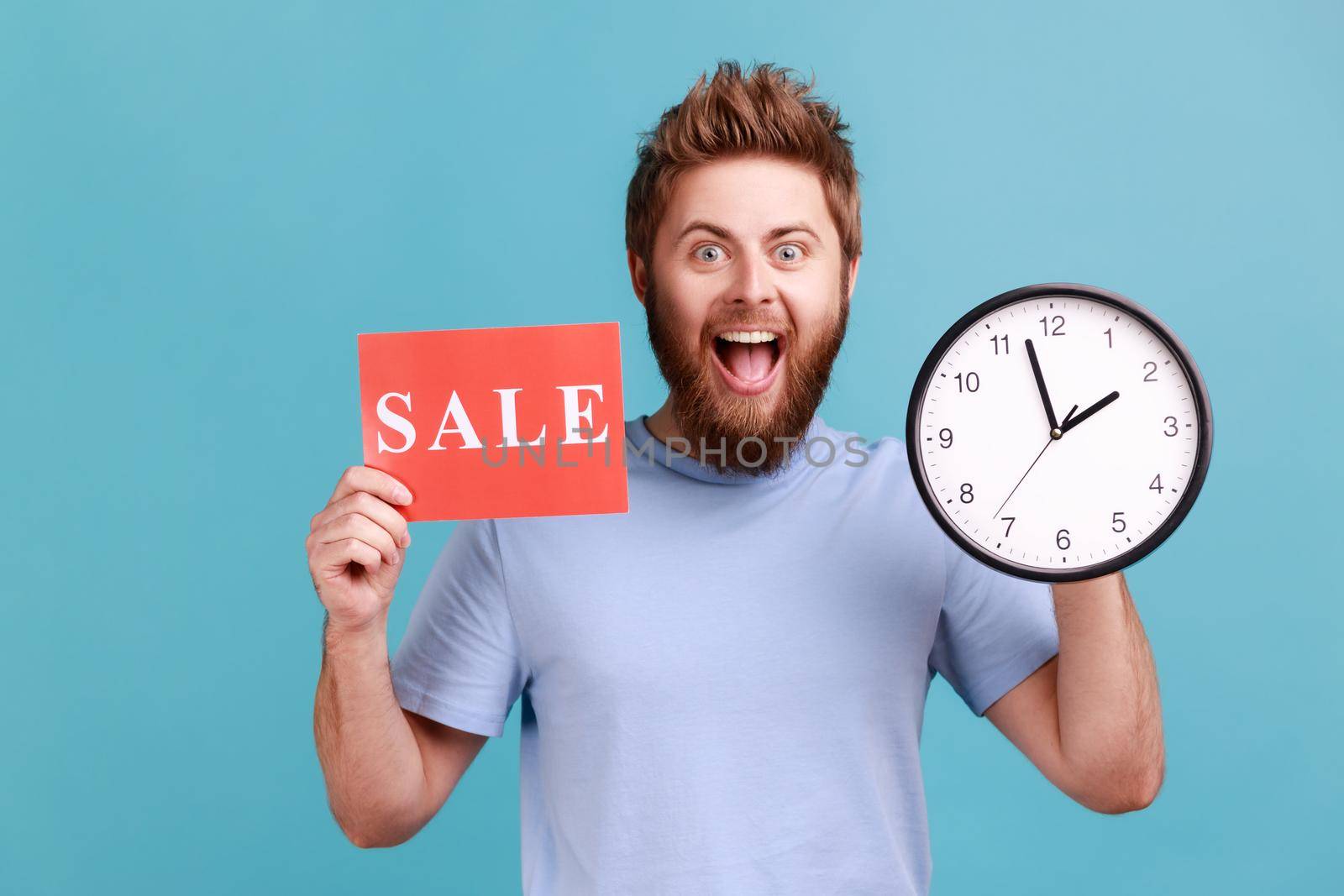 Portrait of excited positive handsome bearded man holding clock and card with sale inscription, announcing finishing huge discounts. Indoor studio shot isolated on blue background.