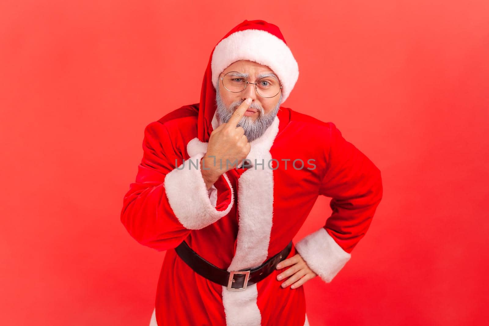 Elderly man with gray beard wearing santa claus costume touching nose, gesturing you are liar, being distrustful of talk, suspecting falsehood. Indoor studio shot isolated on red background.