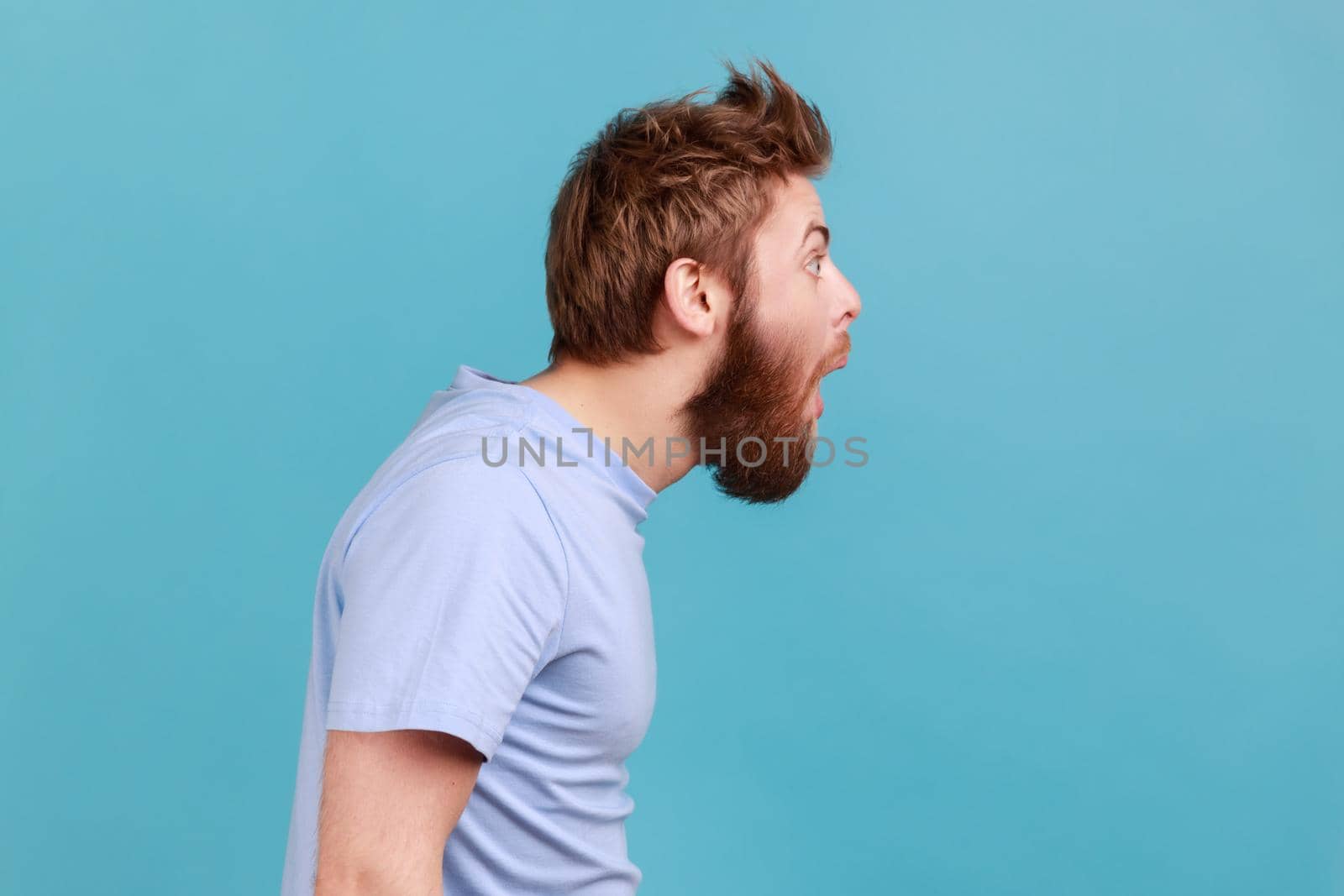 Side view of shocked bearded man gasps from wonder, holds breath, stares at something horrible keeps mouth opened, being astonished. Indoor studio shot isolated on blue background.