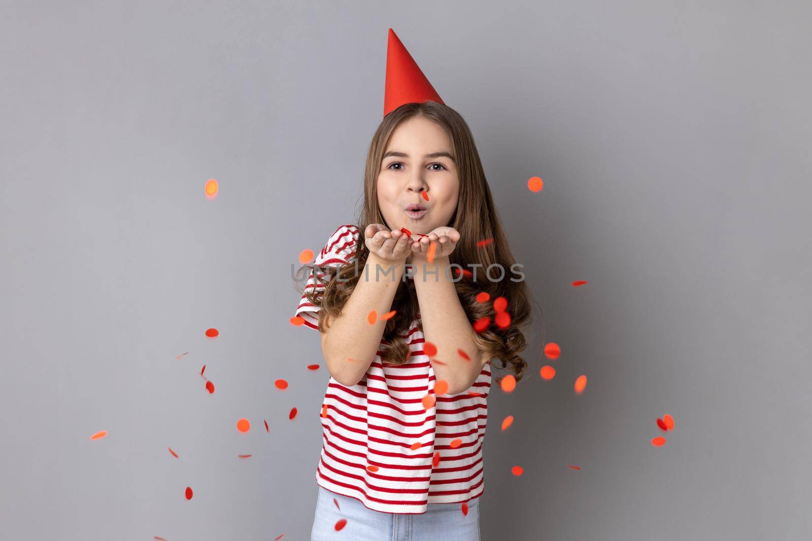 Portrait of happy little girl wearing striped T-shirt and party cone blowing heart shaped confetti, enjoying birthday, festive mood. Indoor studio shot isolated on gray background.