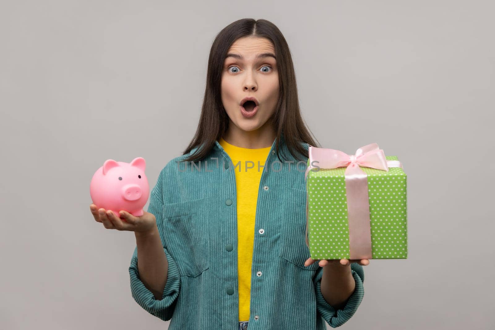 Portrait of amazed surprised woman holding wrapped gift box and piggy bank, save money, looking at camera with open mouth, wearing casual style jacket. Indoor studio shot isolated on gray background.