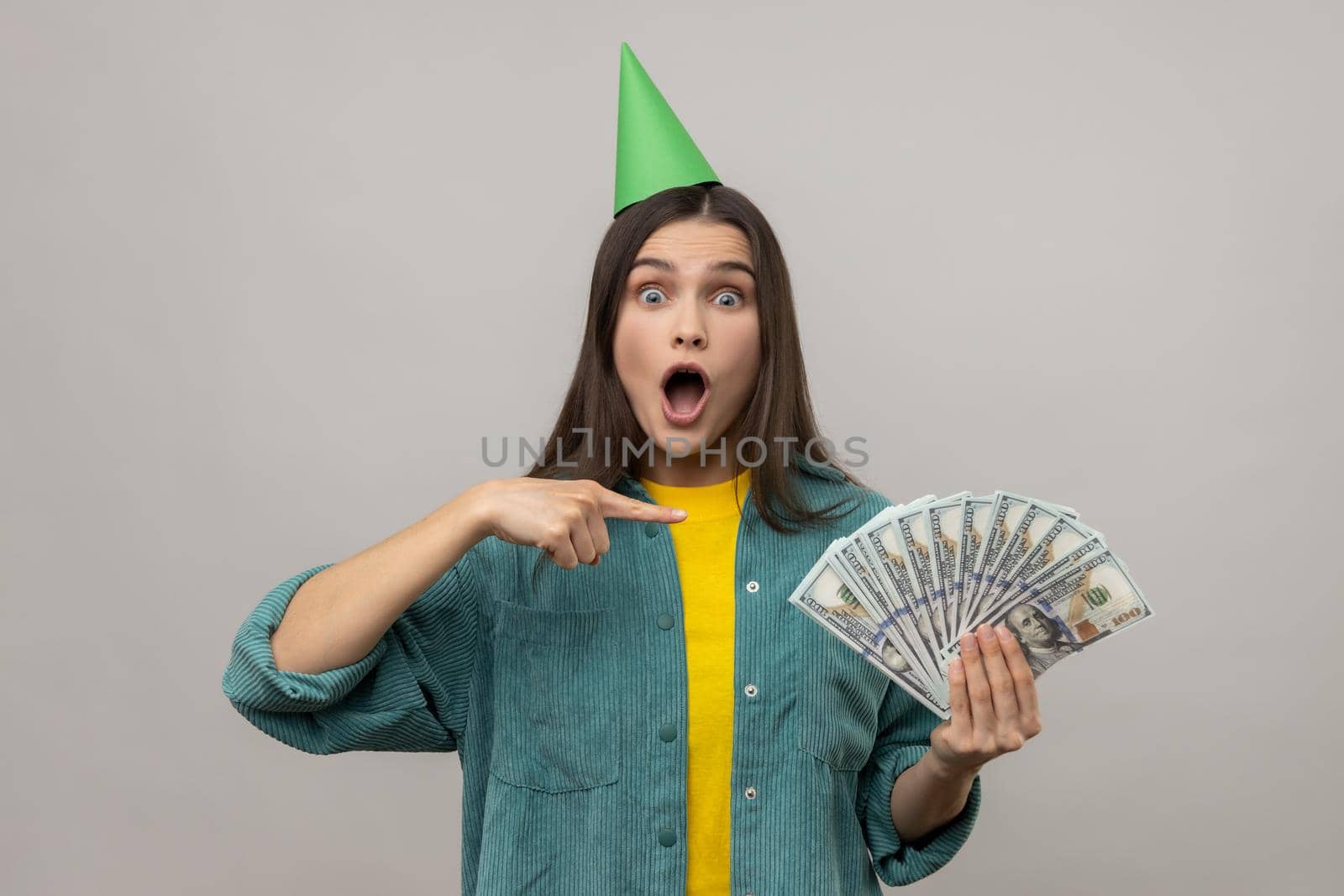 Amazed excited rich woman in party cone on head pointing at money, looking surprised by lottery win, celebrating big profit, wearing casual style jacket. Indoor studio shot isolated on gray background