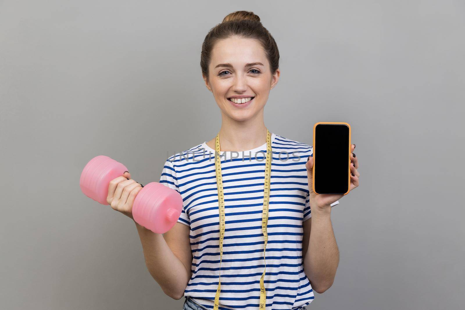 Portrait of smiling happy woman wearing striped T-shirt holding pink dumbbell and mobile phone with empty display for advertisement. Indoor studio shot isolated on gray background.
