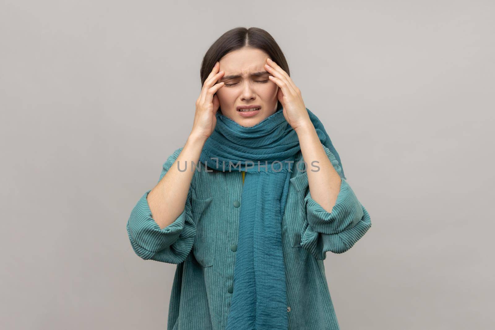 Upset frustrated woman standing wrapped in scarf, feeling headache, cold symptoms, runny nose, frowning face, wearing casual style jacket. Indoor studio shot isolated on gray background.