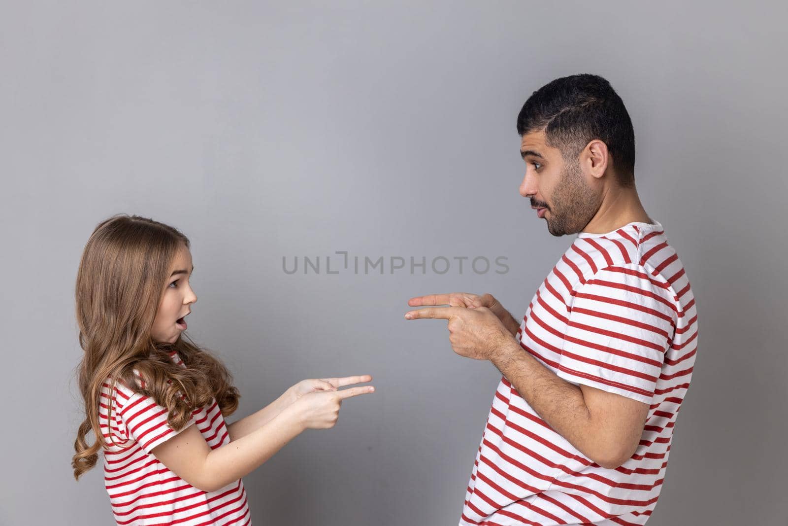 Portrait of surprised astonished father and daughter in striped T-shirts standing pointing at each other, looking with big eyes, choosing turn. Indoor studio shot isolated on gray background.