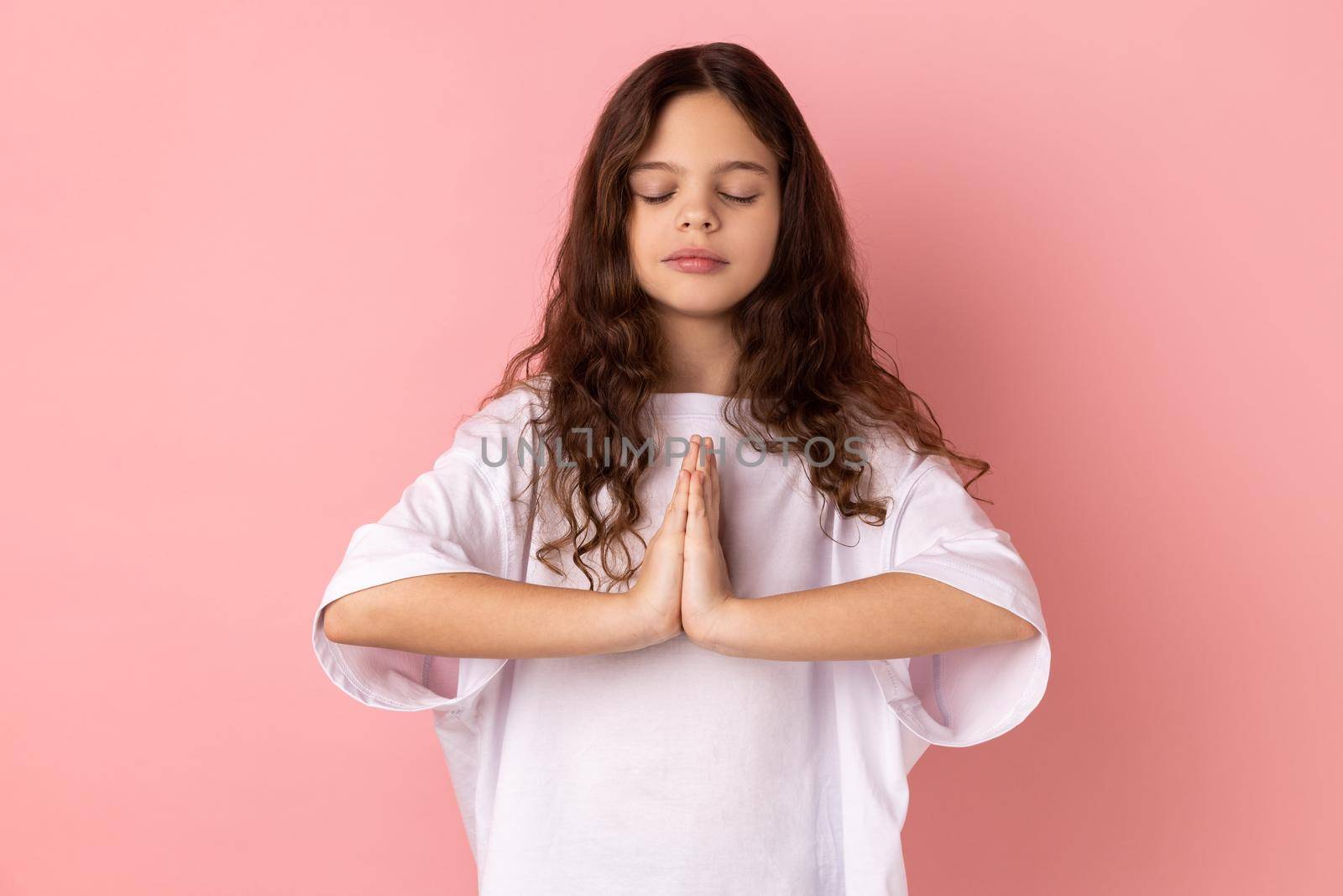 Portrait of little girl wearing white T-shirt keeps hands in yoga gesture, has calm facial expression, keeping palms pressed together and closed eyes. Indoor studio shot isolated on pink background.