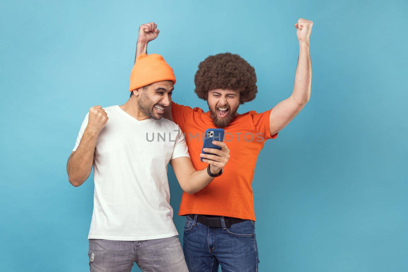 Portrait of two cheerful extremely happy young adult hipster men clenched fists while using mobile phone, screaming yes, betting and winning. Indoor studio shot isolated on blue background.