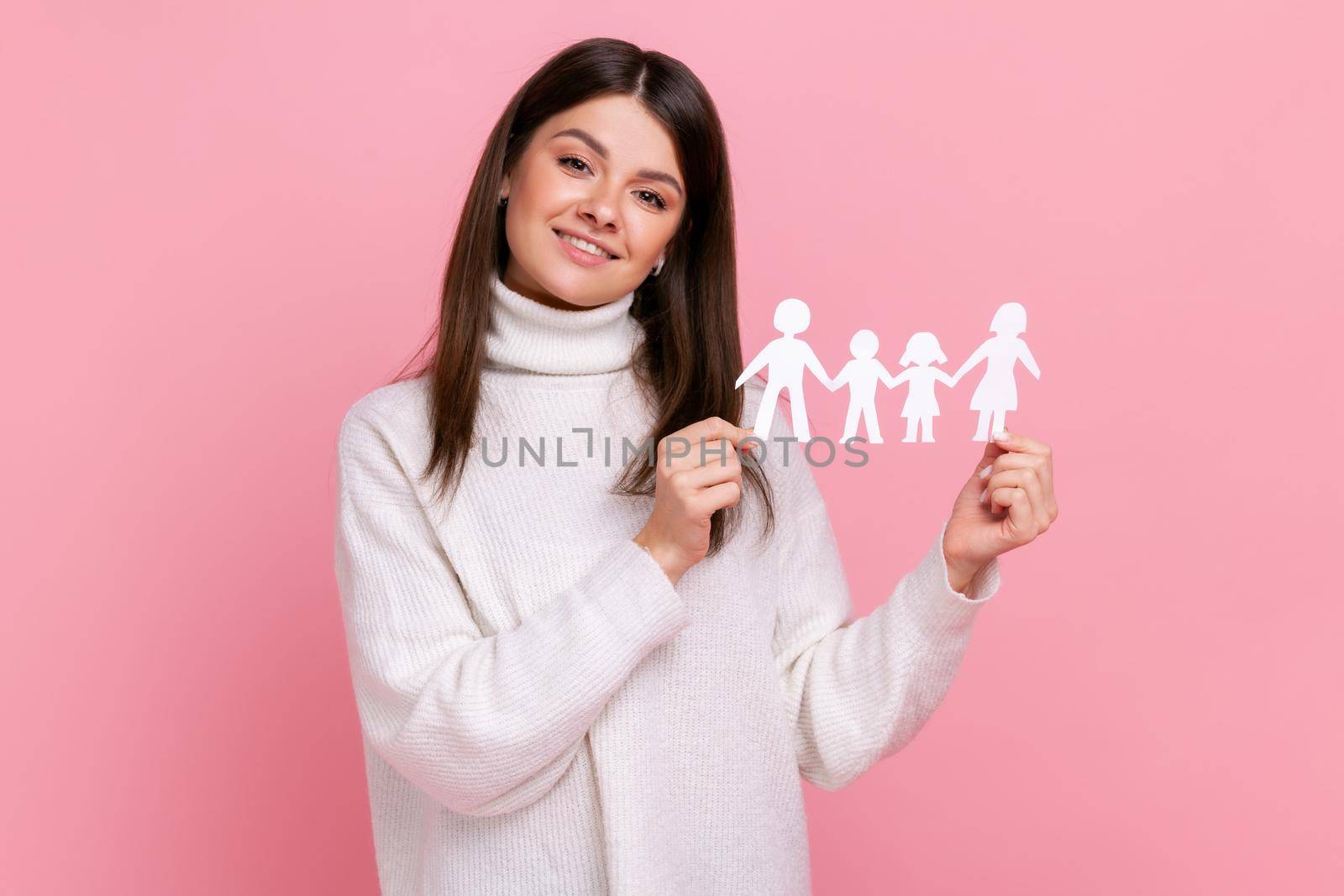 Smiling positive girl holds white paper family chain, looking at camera with happy facial expression, wearing white casual style sweater. Indoor studio shot isolated on pink background.