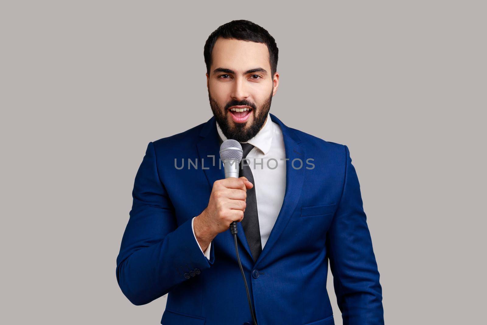 Positive handsome bearded man standing with microphone in hands, sing songs with happy optimistic expression, wearing official style suit. Indoor studio shot isolated on gray background.