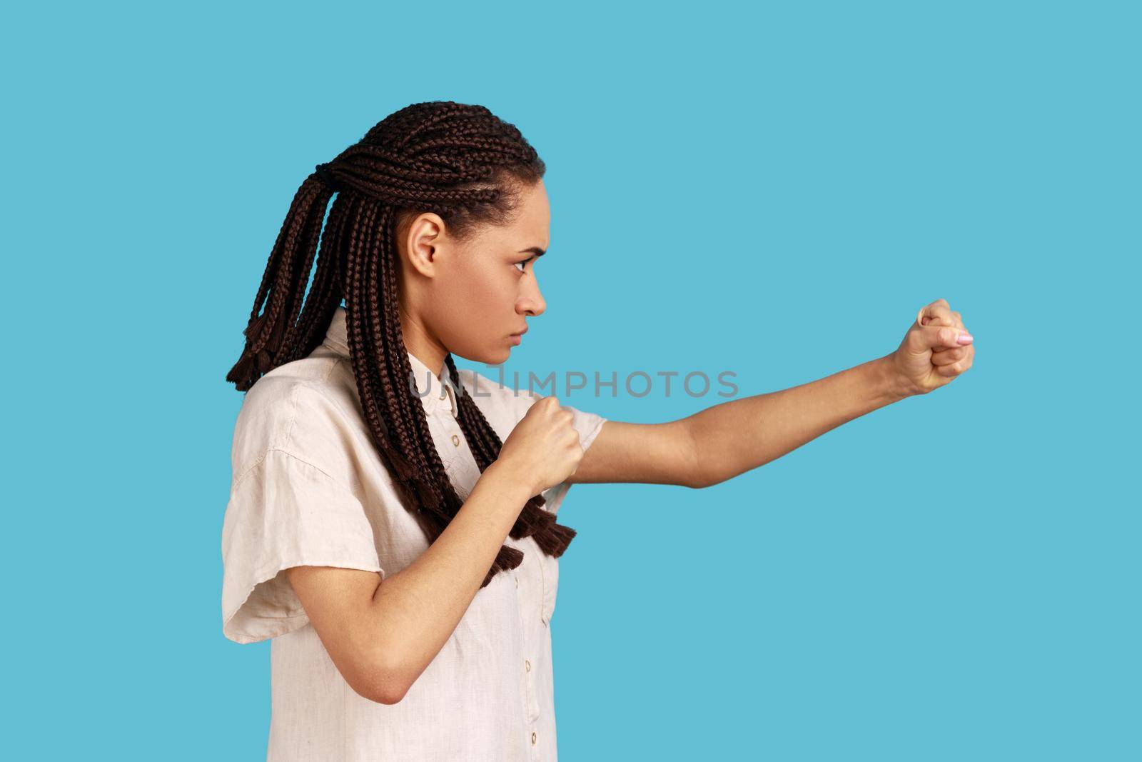 Side view portrait of woman with black dreadlocks having serious look, keeping fists clenched, defencing herself from offence and bad treatment. Indoor studio shot isolated on blue background.