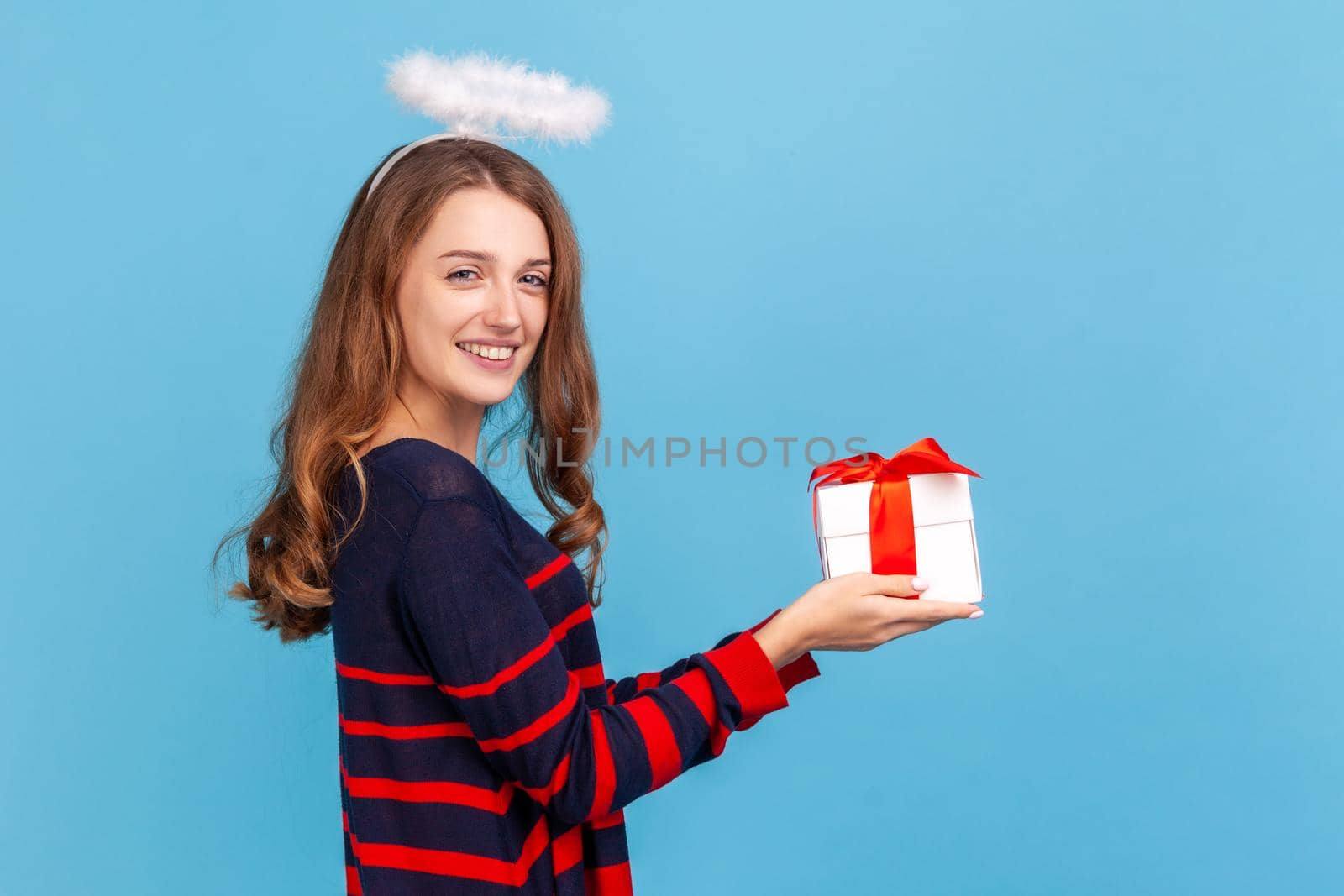 Happy woman wearing striped casual style sweater and nimb over head, giving present box, smiling, congratulating on holiday, looking at camera. Indoor studio shot isolated on blue background.
