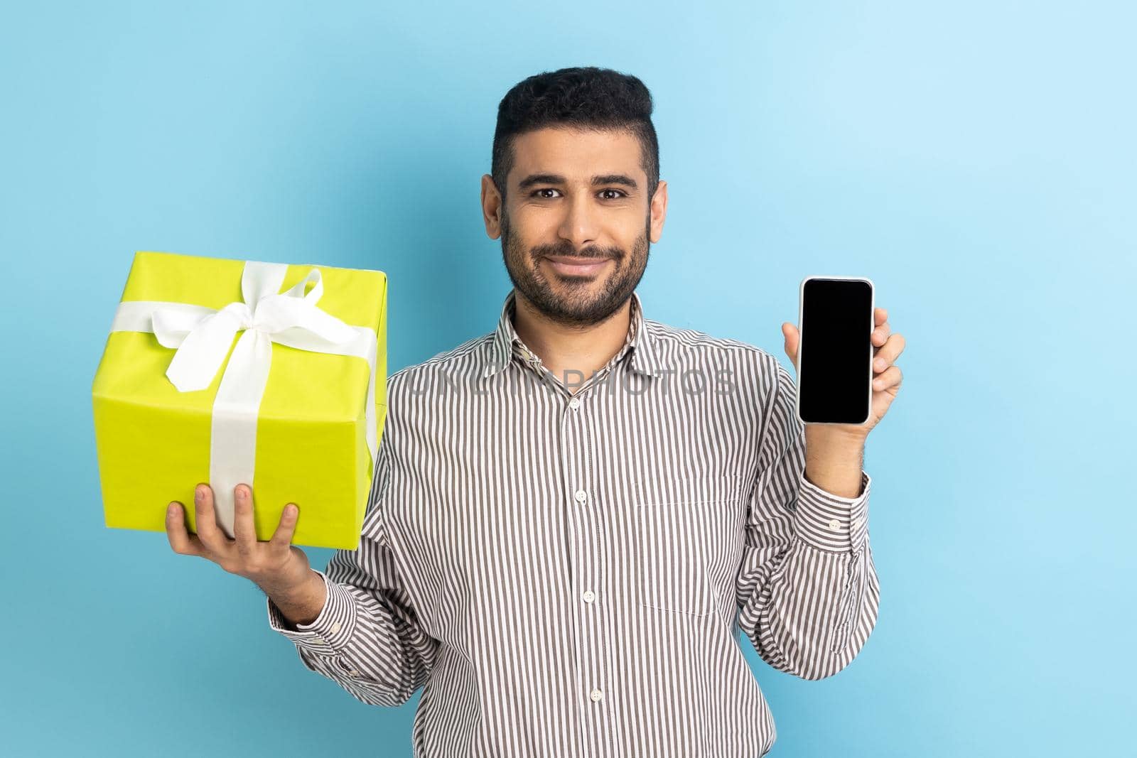 Happy bearded businessman holding present box and smatrphone with blank screen for promotion, looking smiling at camera, wearing striped shirt. Indoor studio shot isolated on blue background.