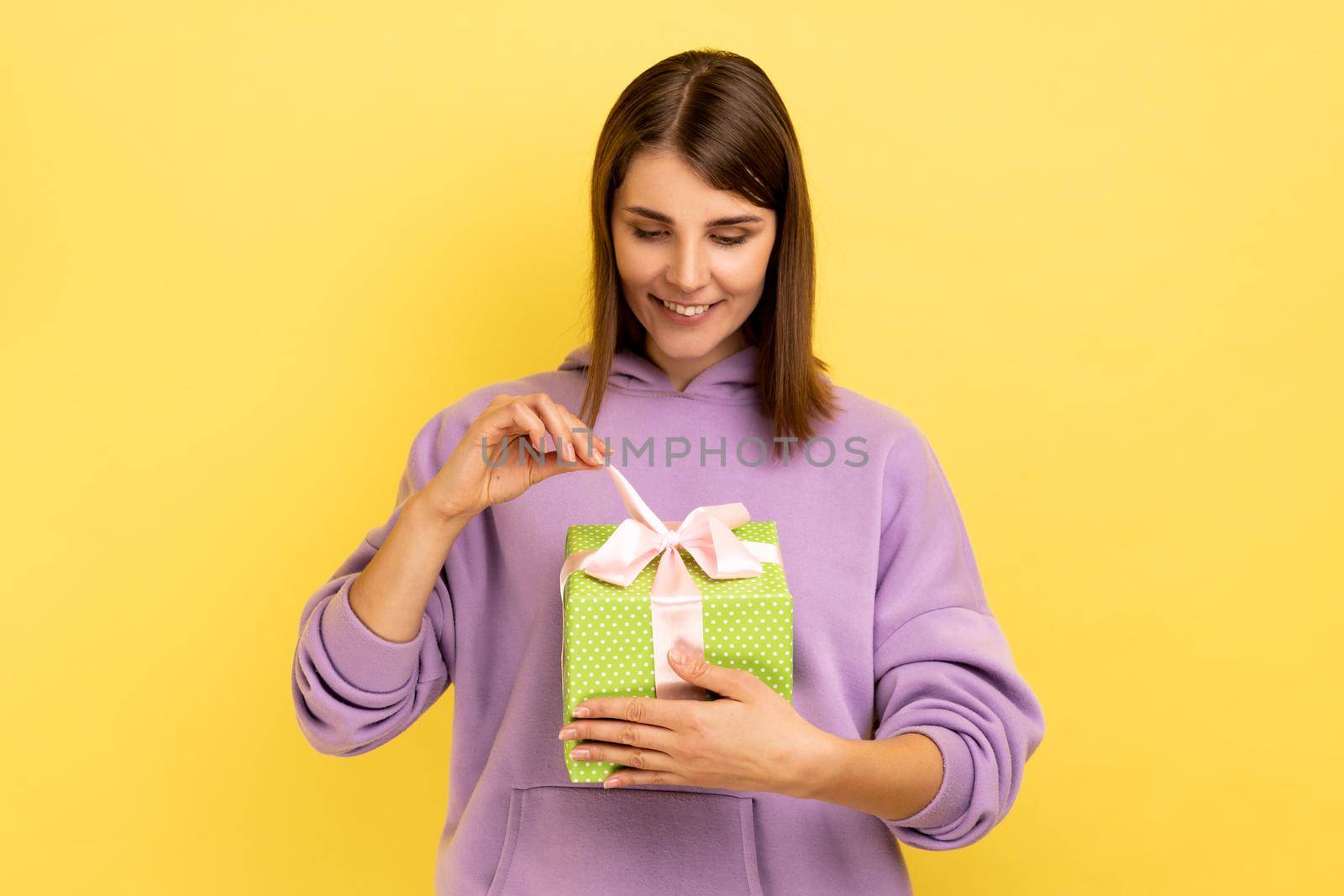Young beautiful smiling woman feeling happy, opens present box with happy satisfied expression, being glad to get gift, wearing purple hoodie. Indoor studio shot isolated on yellow background.