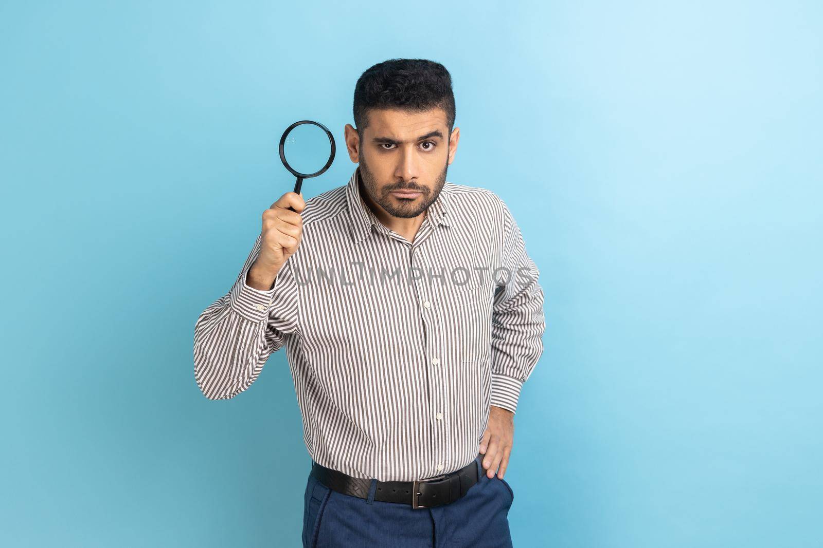Serious man with beard looking at camera and holding magnifying glass, spying, finding out something, exploring, inspecting, wearing striped shirt. Indoor studio shot isolated on blue background.