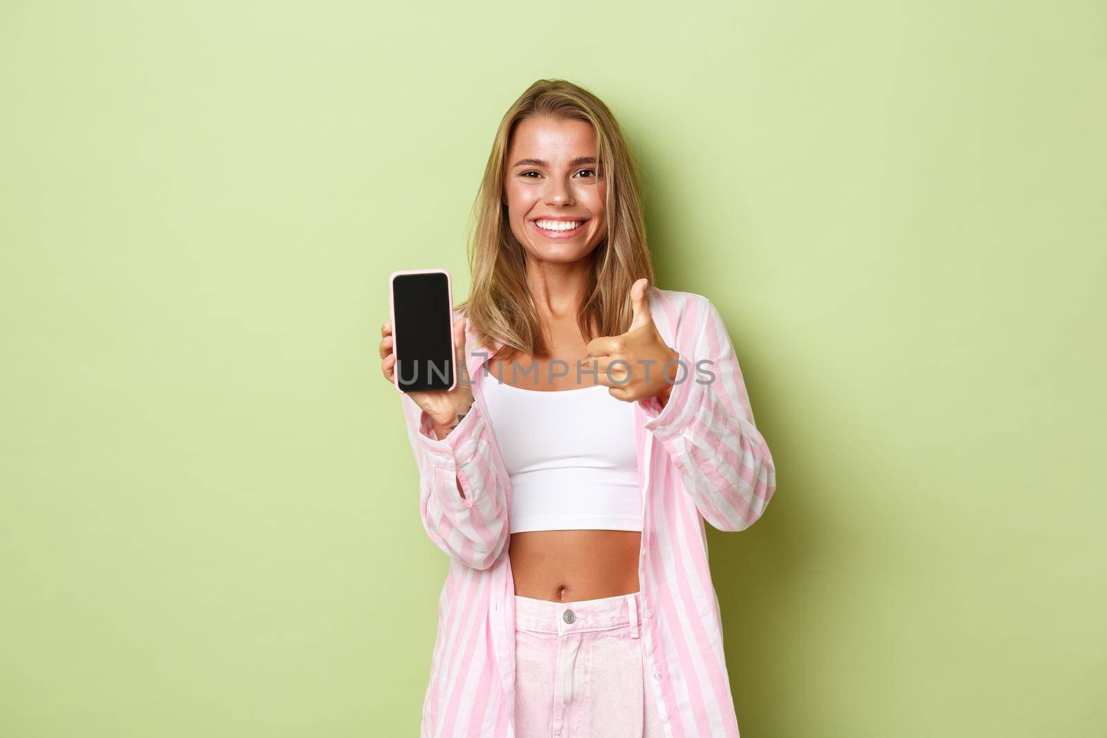 Sassy beautiful blond woman in pink shirt, showing something good on smartphone screen, make thumbs-up in approval and smiling, standing over green background by Benzoix