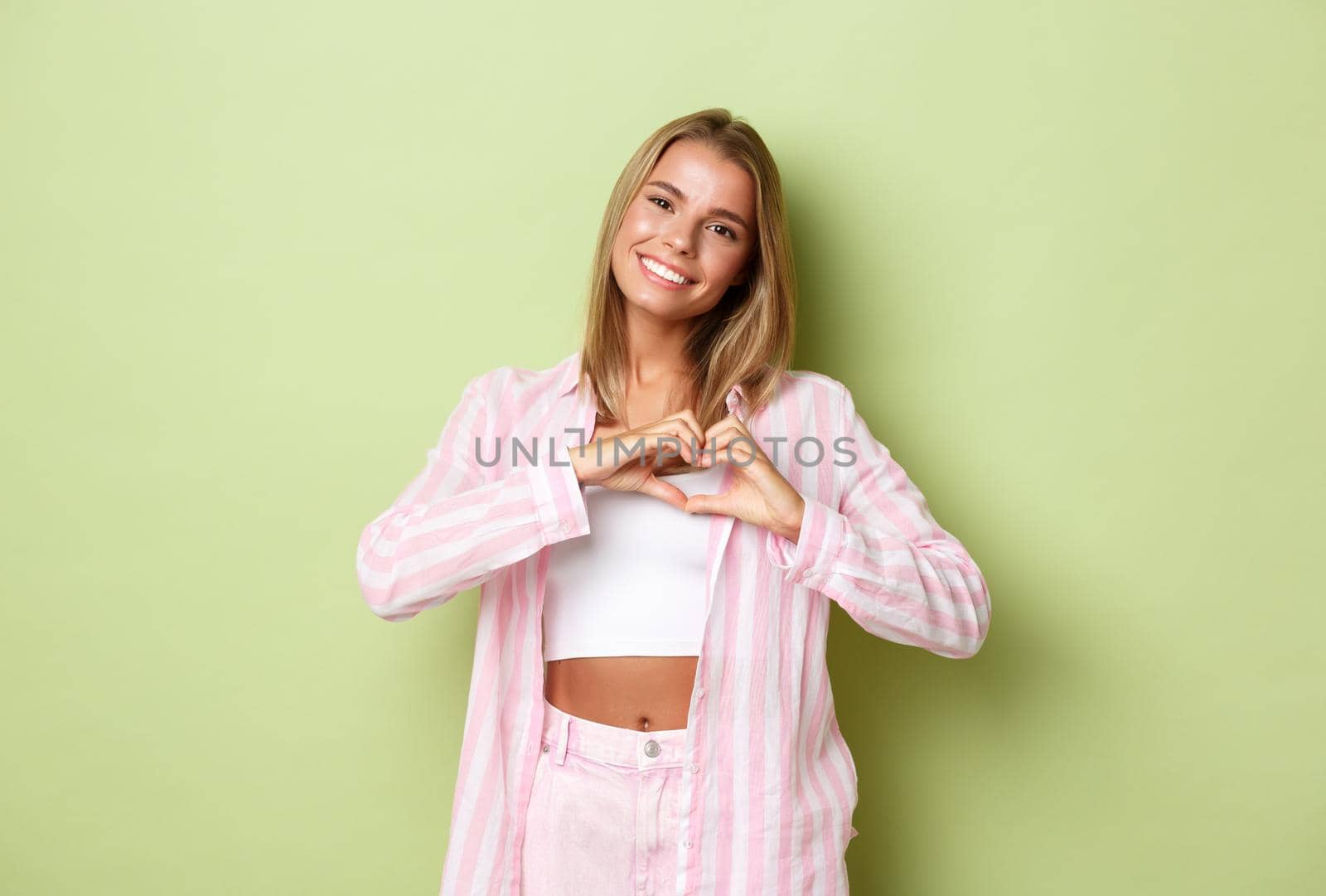 Portrait of beautiful lovely woman with blond short hairstyle, wearing pink outfit, showing heart sign and smiling, confess in love or sympathy, standing over green background by Benzoix