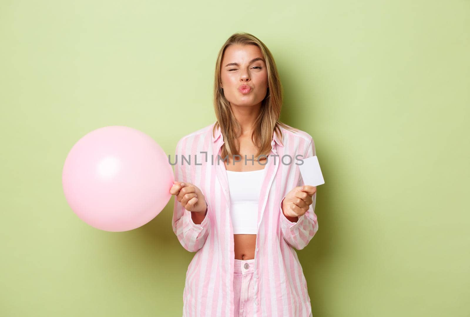 Portrait of attractive blond girl in glamour outfit, holding balloon and credit card, standing over green background.