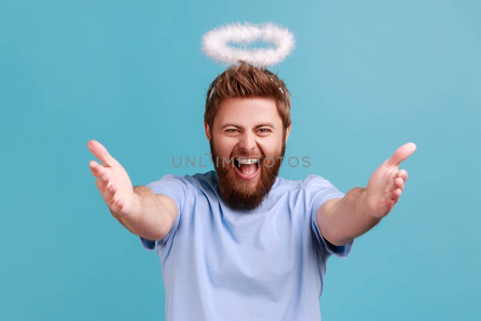 Portrait of excited angelic man with holy nimbus on head standing opening his hands wide to hug, welcoming with friendly smile, sharing love. Indoor studio shot isolated on blue background.