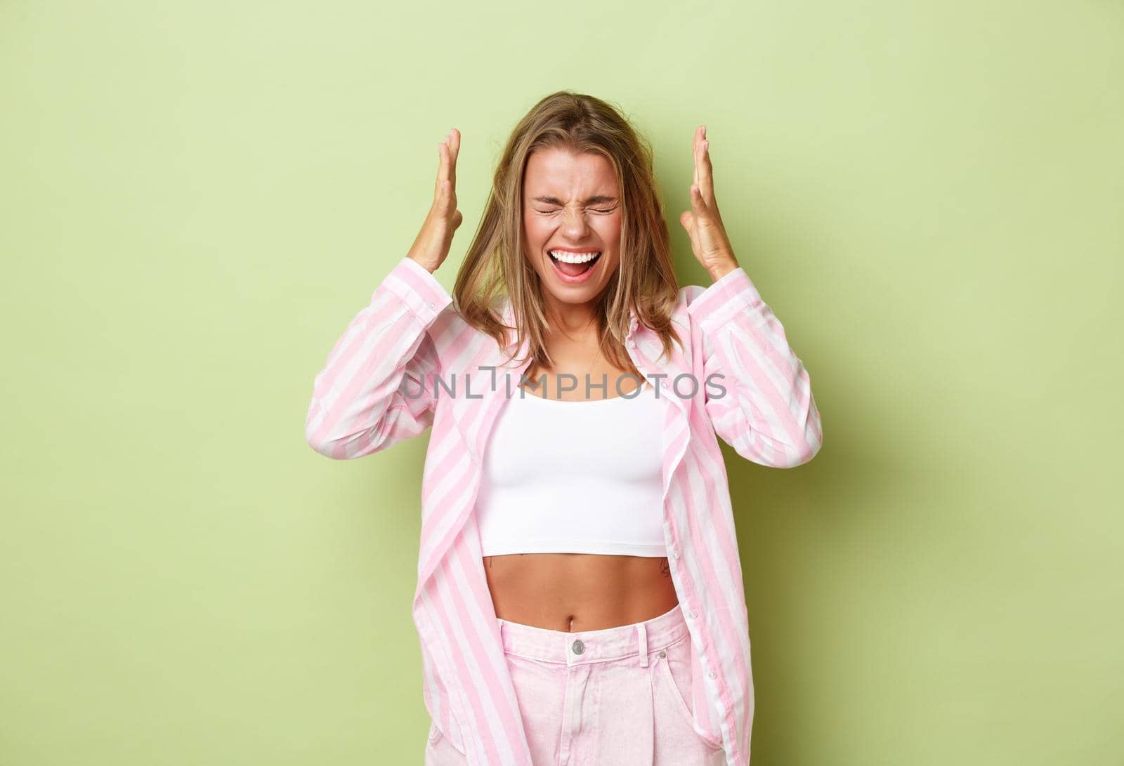 Potrait of distressed blond woman in pink shirt, holding hands on head and screaming, having depression, standing over green background.