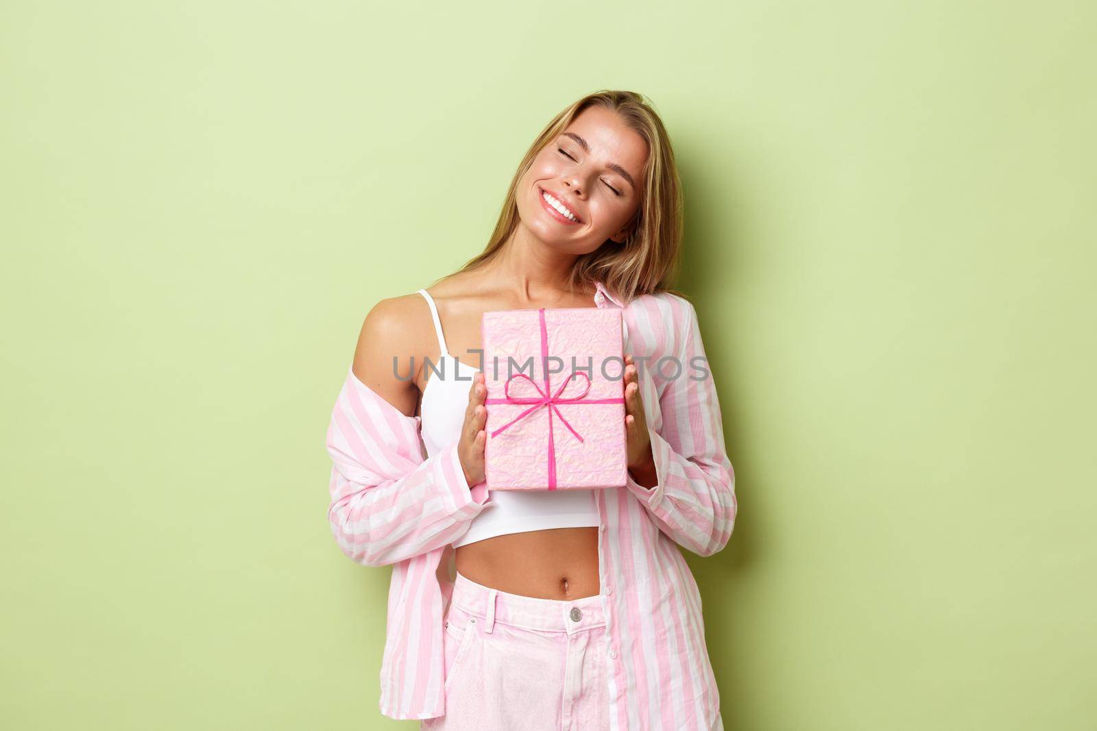 Close-up of dreamy beautiful woman with blond hair,smiling delighted, receiving a gift, standing over green background by Benzoix